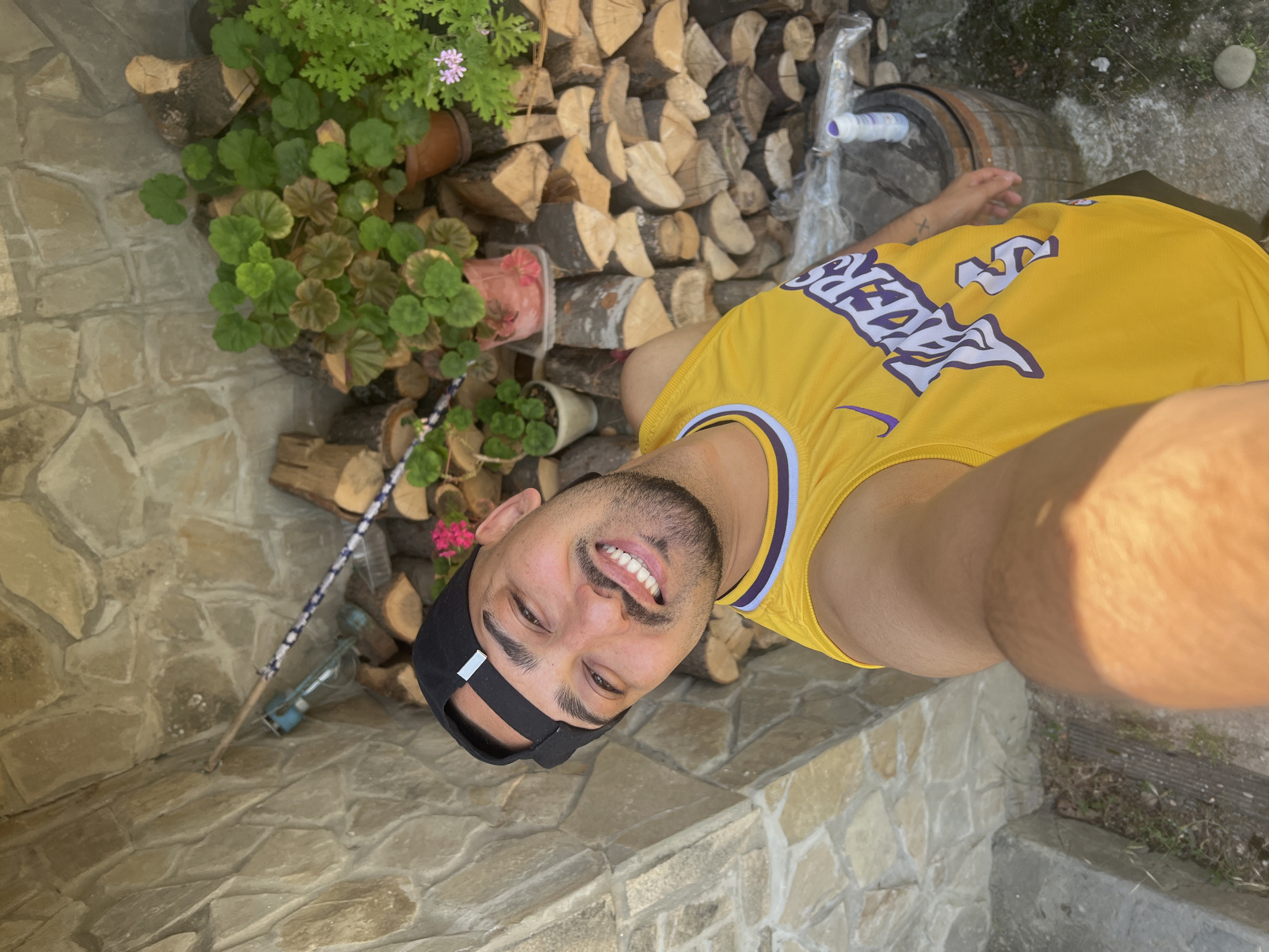 A man wearing a yellow Los Angeles Lakers jersey and a black baseball cap is taking a selfie outdoors. He has a beard and is smiling. Behind him, there is a stone wall, a stack of firewood, potted plants, and a wooden barrel. The location is identified as the city of Shumata, Bulgaria. The scene appears to be a rustic outdoor setting, possibly a yard or a garden.