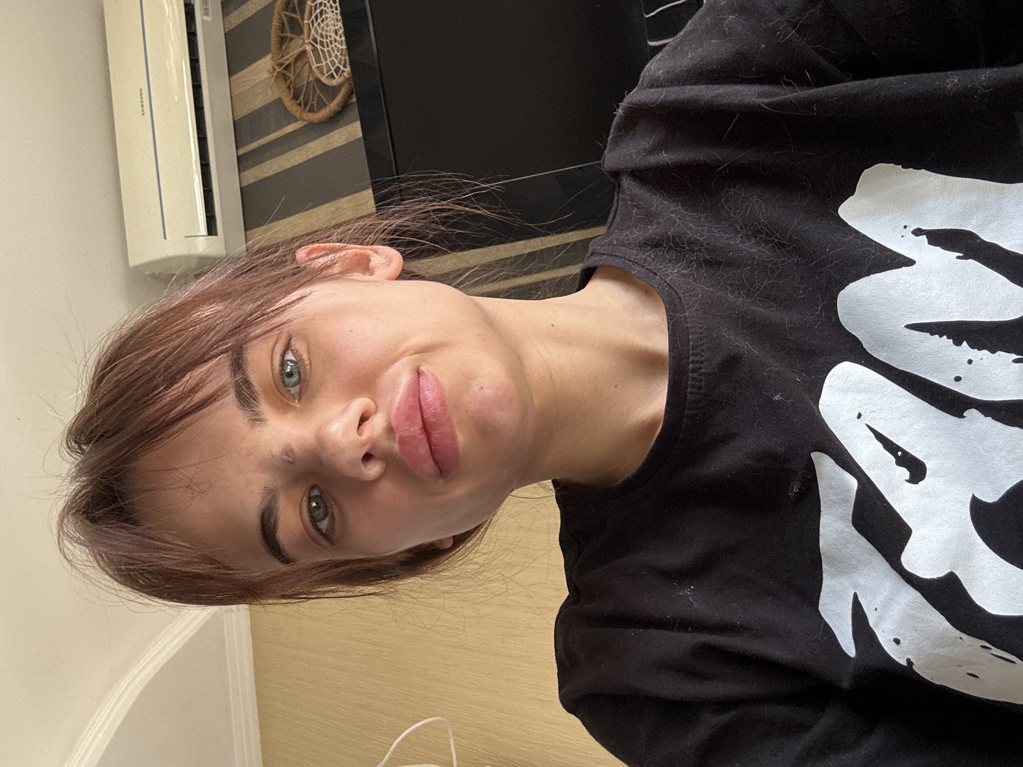 A close-up, slightly angled portrait of a woman with light brown hair and blue eyes. She is wearing a black t-shirt with a large white design on the front. The background includes a light-colored wall, an air conditioner, and a decorative woven item. The location in the city of Rostov-on-Don, Russia, is not apparent from the image.