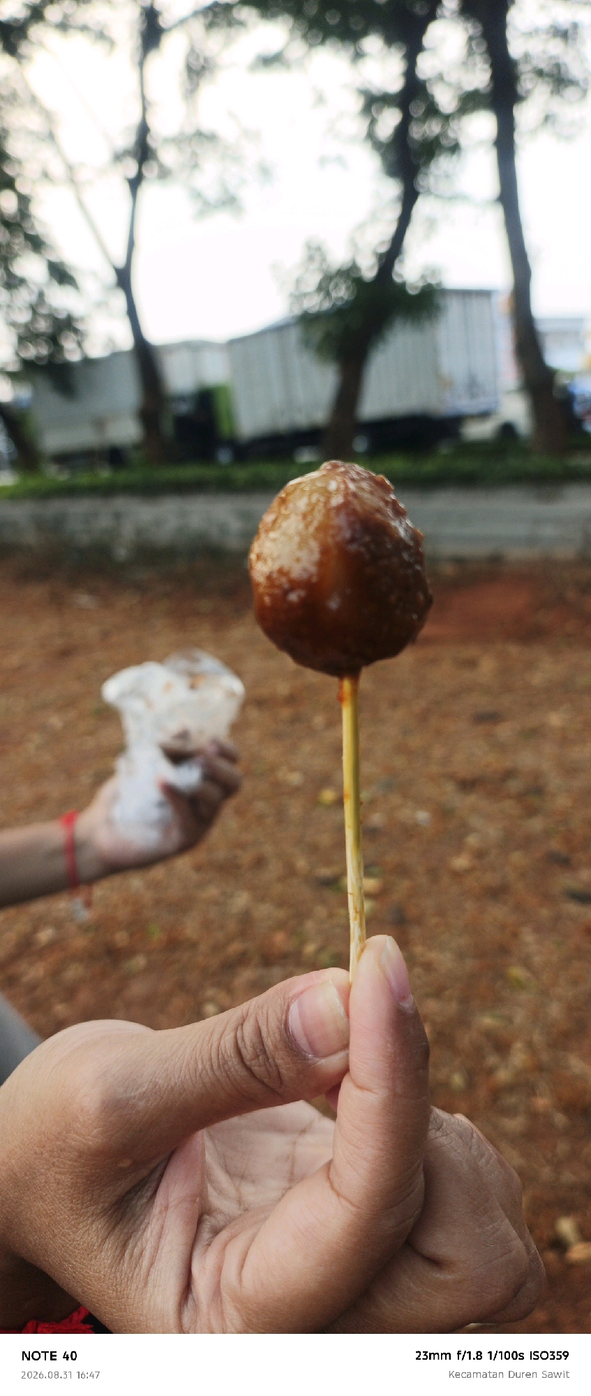 In this close-up shot, a person's hand holds a single meatball on a skewer. The meatball, coated in a glistening sauce, is the primary subject, positioned slightly off-center. The background is blurred, showing trees and what appears to be a white truck, suggesting an outdoor setting. The lower portion of the image displays text that reads "NOTE 40", "2026.08.31 16:47", "23mm f/1.8 1/100s ISO359", and "Kecamatan Duren Sawit". The metadata indicates the photo was taken in Kecamatan Duren Sawit, a subdistrict within East Jakarta, Indonesia.