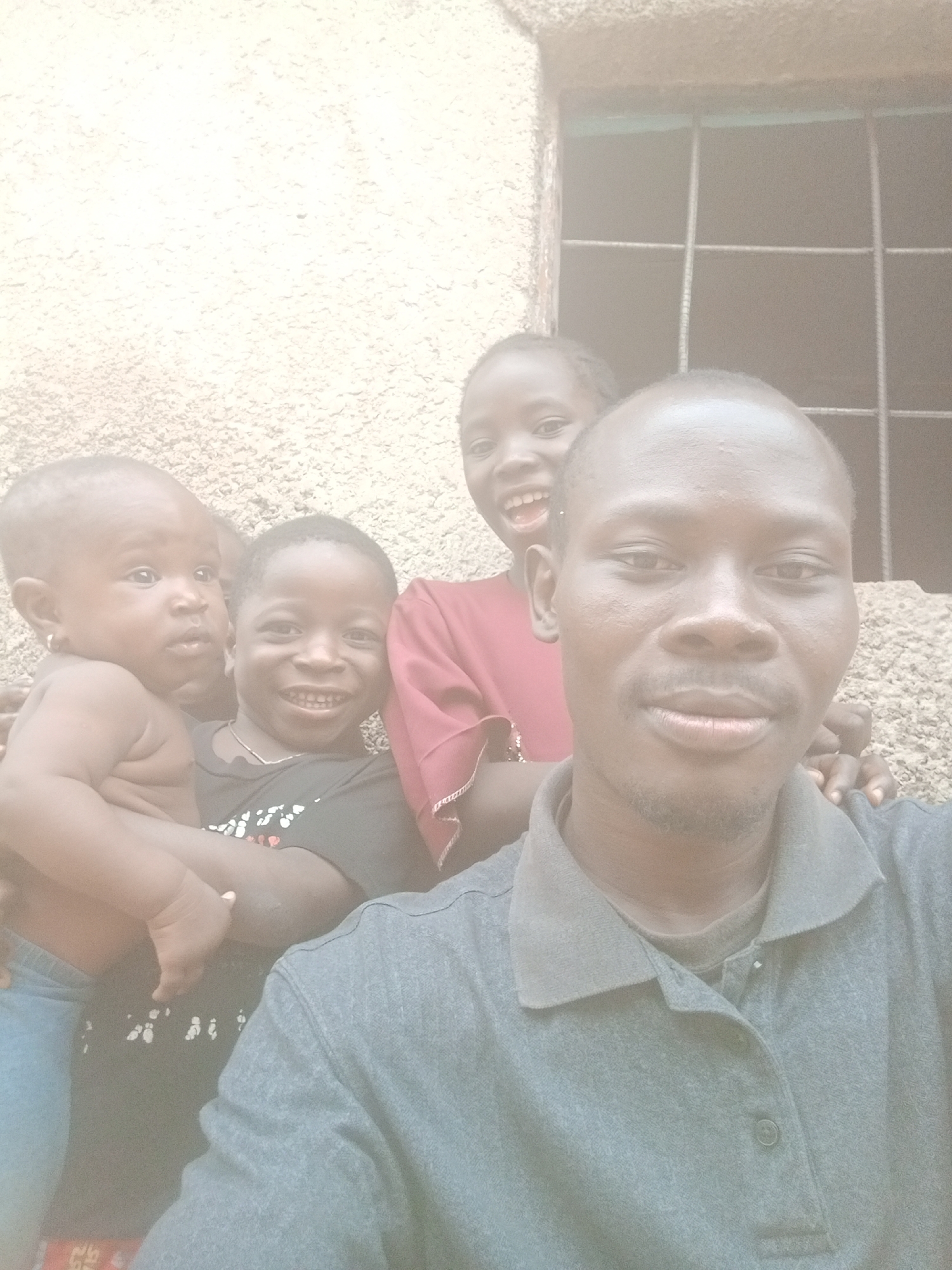 A medium shot captures a man and three children posing in front of a textured concrete wall and a window with a metal grid. The man is in the foreground, wearing a gray collared shirt, and has a short haircut and a neatly trimmed mustache and goatee. To the left of the man, a baby is being held, its bare skin contrasting with its denim pants. Beside the baby, a young boy is seen wearing a dark t-shirt with white designs and a silver necklace, his face lit up with a broad smile. In the background, a young girl in a red shirt is visible, her mouth open in a wide grin. The concrete wall behind them is rough, and a dimly lit window with a grid pattern is situated on the upper right. The location is identified as Sabon Tasha, Nigeria.