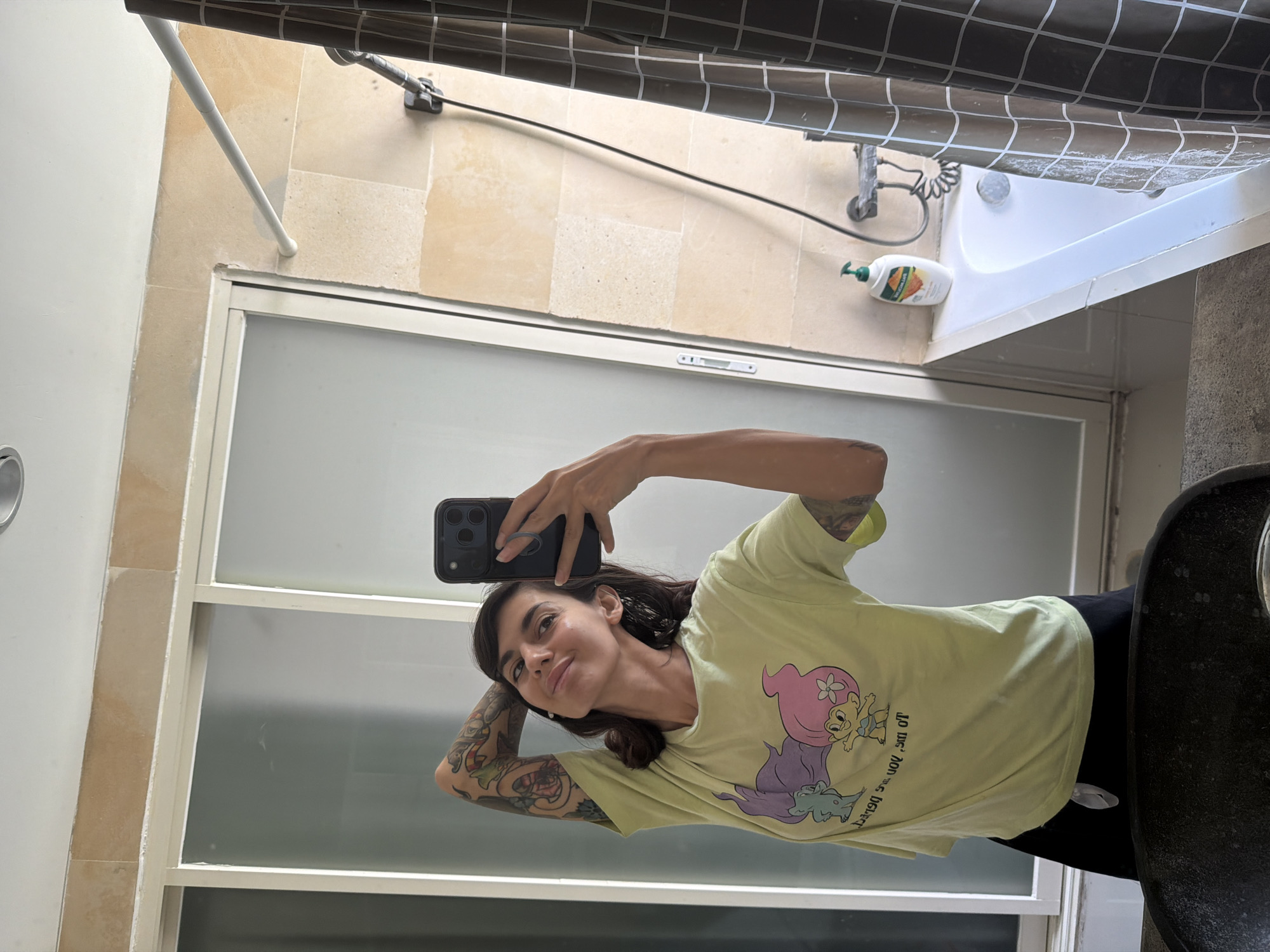 In Kuta Selatan, Indonesia, a woman with dark hair and tattoos on her arms poses for a mirror selfie in a bathroom. She is wearing a light green t-shirt with a cartoon graphic and black shorts. Her right arm is bent, with her hand behind her head, and she is holding a black smartphone in her left hand. The bathroom features beige tiled walls, a sliding glass door, a white bathtub, a showerhead, and a brown checkered shower curtain.