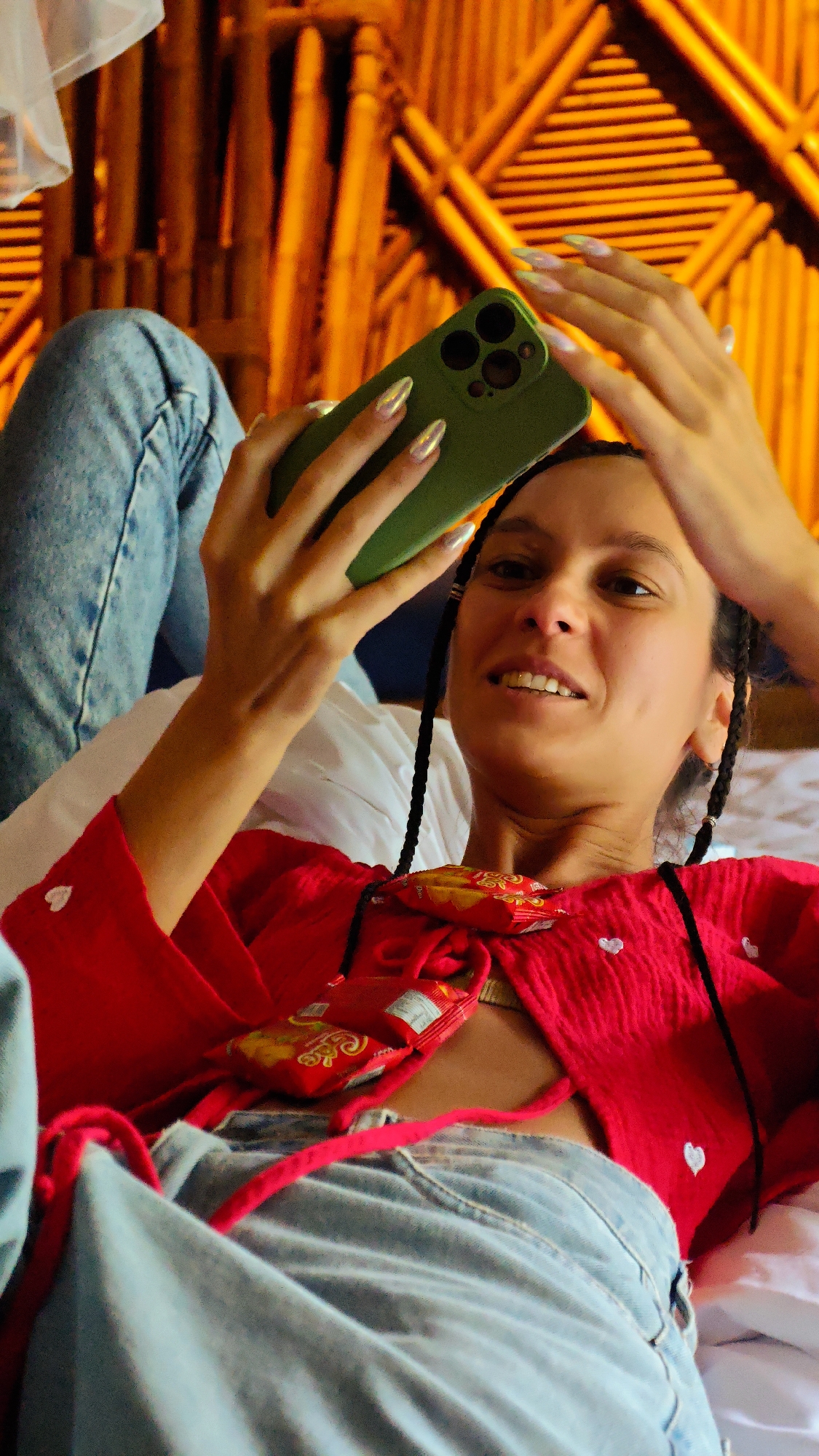 A young adult female is depicted lying on her back on white bedding, holding a dark green smartphone with a protective case above her head, looking directly towards the viewer with a slight smile. Her dark hair is styled in multiple thin braids. She wears a red long-sleeved top with small white heart-shaped embellishments and light blue denim jeans. The red top is partially open, revealing a light-colored garment underneath, and a red packaged snack (appearing to be cookies or biscuits, with "CHOCO" and "Oreo" text visible alongside Vietnamese script) is tucked into the neckline. Her fingernails are long, pointed, and have a metallic, iridescent finish. The background consists of a wall or headboard constructed from horizontal bamboo or wooden poles, illuminated with a warm orange light. The scene is located in Mu Cang Chai, Vietnam.