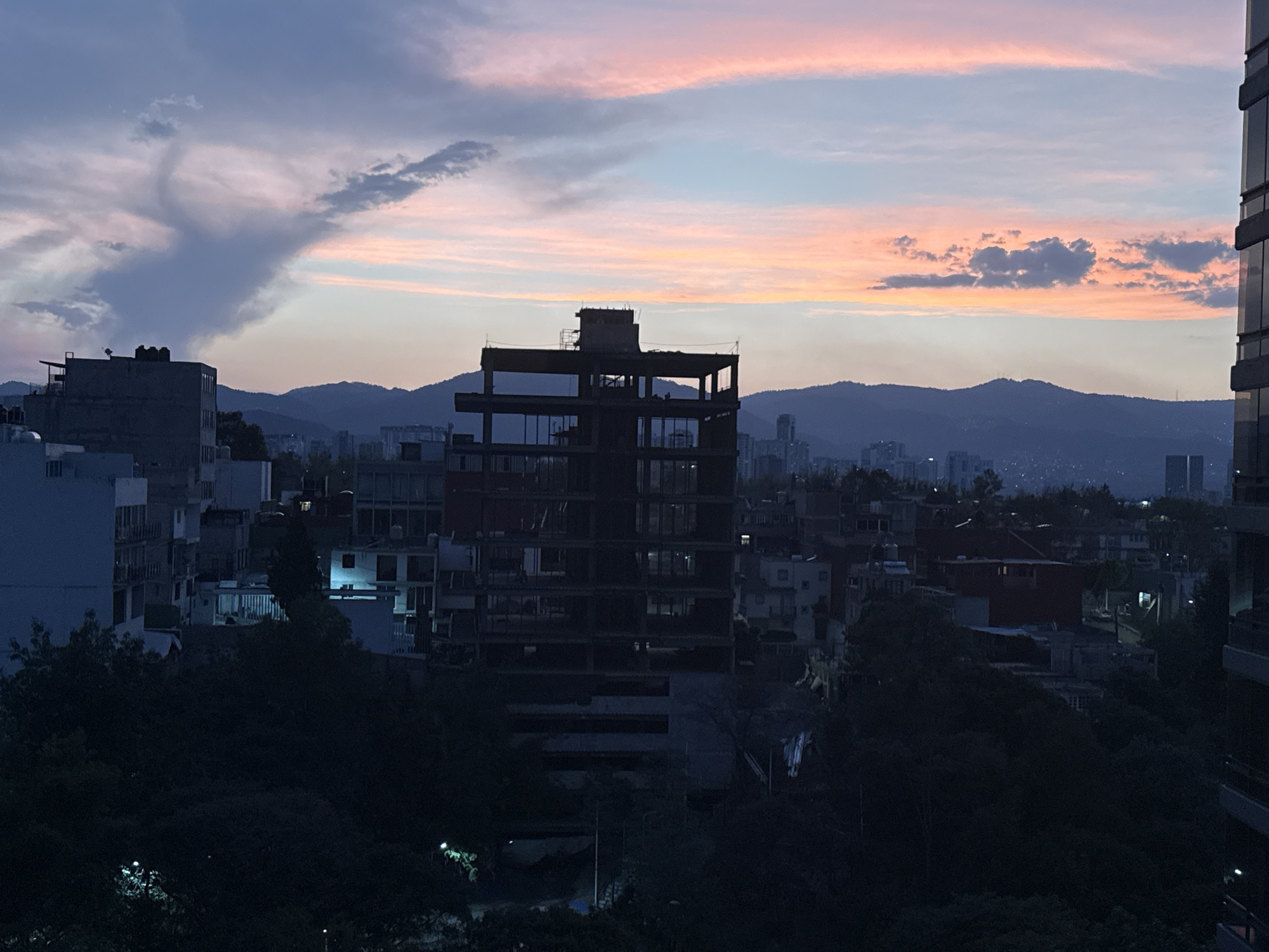 This image shows a view of Mexico City at dusk. In the foreground, there is an unfinished, multi-story building under construction. Behind the unfinished building are other buildings of varying heights, typical of an urban landscape. In the distance, a mountain range forms the horizon. The sky is a mix of blue, gray, and pink hues, indicating the transition from day to night. The lighting suggests the image was taken shortly before nightfall, as the buildings and trees in the foreground are largely silhouetted.