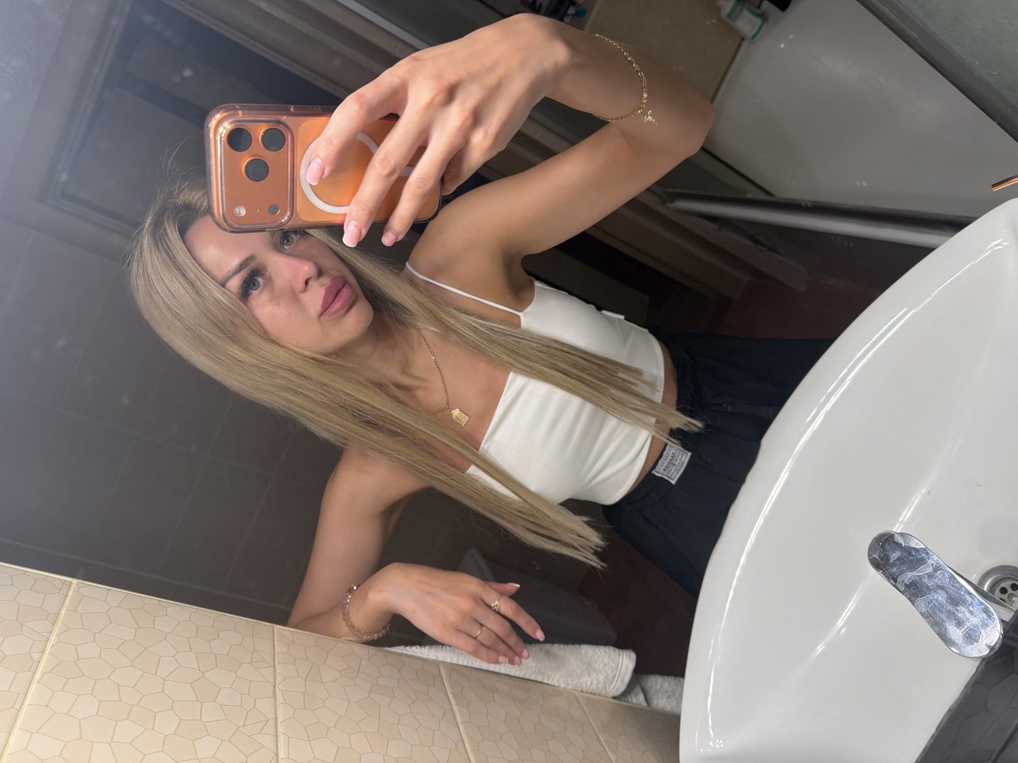 The image is a mirror selfie taken in a bathroom. A young woman with long blonde hair is reflected in the mirror, holding an orange smartphone. She is wearing a white crop top and black pants. Her right arm is extended to take the photo, and her left arm is resting on the edge of the sink. The bathroom appears to be tiled with light-colored hexagonal tiles on the left wall and dark tiles on the right. A white sink with a chrome faucet is visible in the foreground. The background is indistinct due to the mirror reflection, showing what appears to be a doorway and a closet. The overall lighting suggests an indoor environment, likely a home bathroom. The image does not provide any specific indicators of its location within the city of Kyiv, Ukraine.