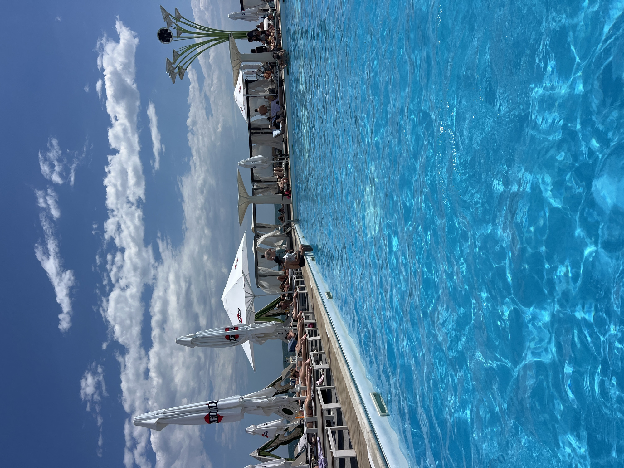 This is a photograph taken at a poolside resort in Fontanka, Ukraine, on a sunny day with a partly cloudy blue sky. The foreground is dominated by a large, vibrant blue swimming pool with clear water, reflecting the sunlight. Along the edge of the pool, there is a wooden deck with numerous lounge chairs. Several people are seen relaxing on these chairs, sunbathing, or sitting up. Some of the chairs are shaded by large, white umbrellas, some of which have "Martini" branding visible. Further back, there are cabanas providing more shade. In the distance, beyond the resort amenities, the ocean can be seen. A distinctive, tree-like sculpture with solar panels is also visible on the right side of the frame.