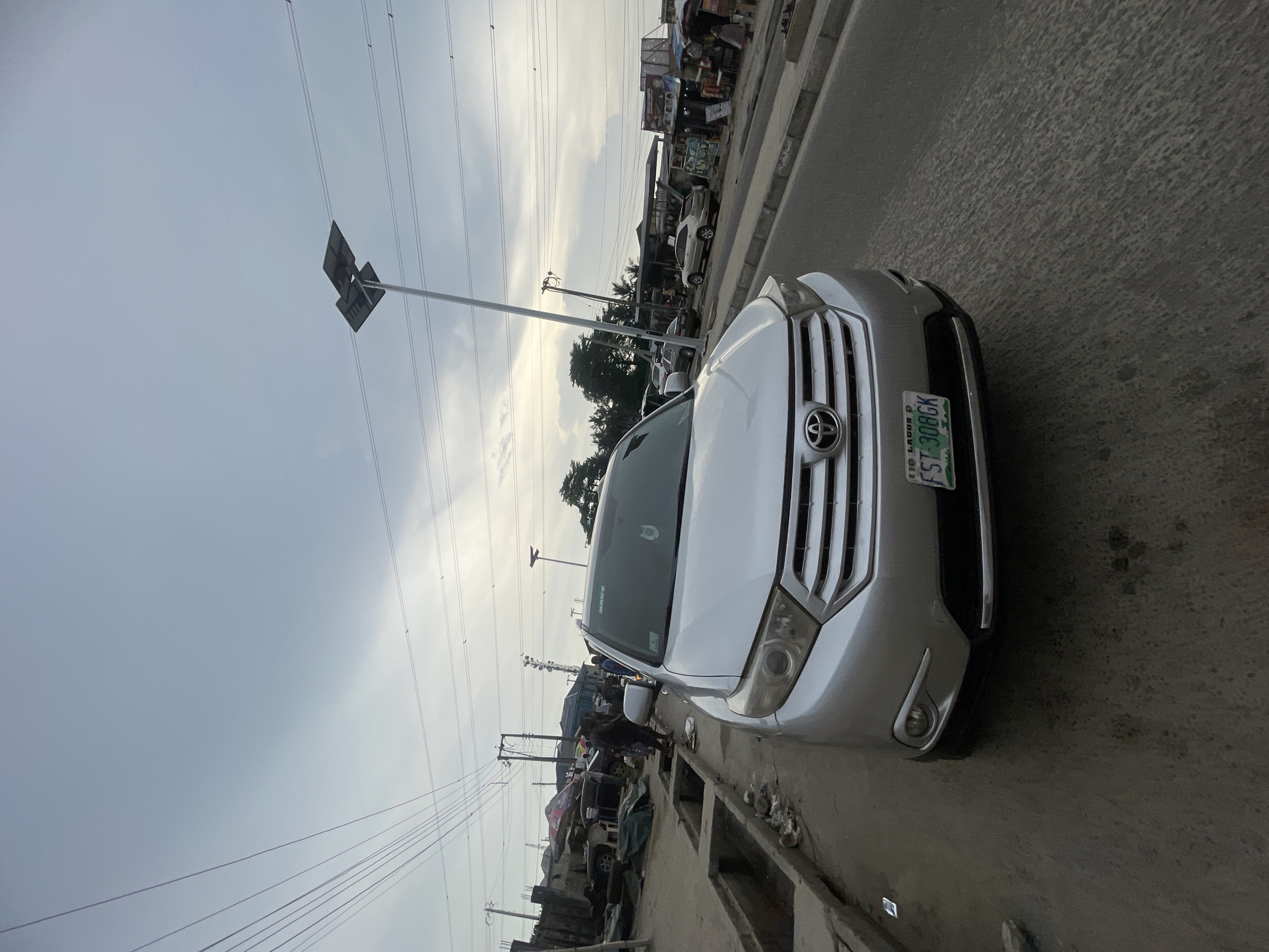 The image captures an outdoor street scene, likely in an urban or semi-urban area, under an overcast sky. A silver or light-colored Toyota Highlander SUV dominates the foreground, parked along the roadside and angled slightly towards the viewer. Its front grille, headlights, and the prominent Toyota emblem are clearly visible.
On the front bumper, a green Nigerian license plate with white lettering is seen, displaying "FST-308GK" and "LAGOS P," indicating its registration in Lagos State.
The street itself is an asphalt road with a textured surface, flanked by a concrete curb and a visible drainage ditch or gutter. Along the side of the road where the SUV is parked, a bustling environment unfolds with numerous makeshift market stalls or small shops crammed together. Several other vehicles are partially visible, parked along this commercial strip.
In the background, a few blurry figures of people can be discerned, engaged in daily activities near the stalls and parked cars. Overhead, a network of power lines stretches across the frame, supported by tall utility poles. One pole features a square sign or light fixture. The sky is predominantly covered with light gray clouds, creating a diffused, natural light. The scene portrays a typical, active streetscape in a Nigerian city or town.
