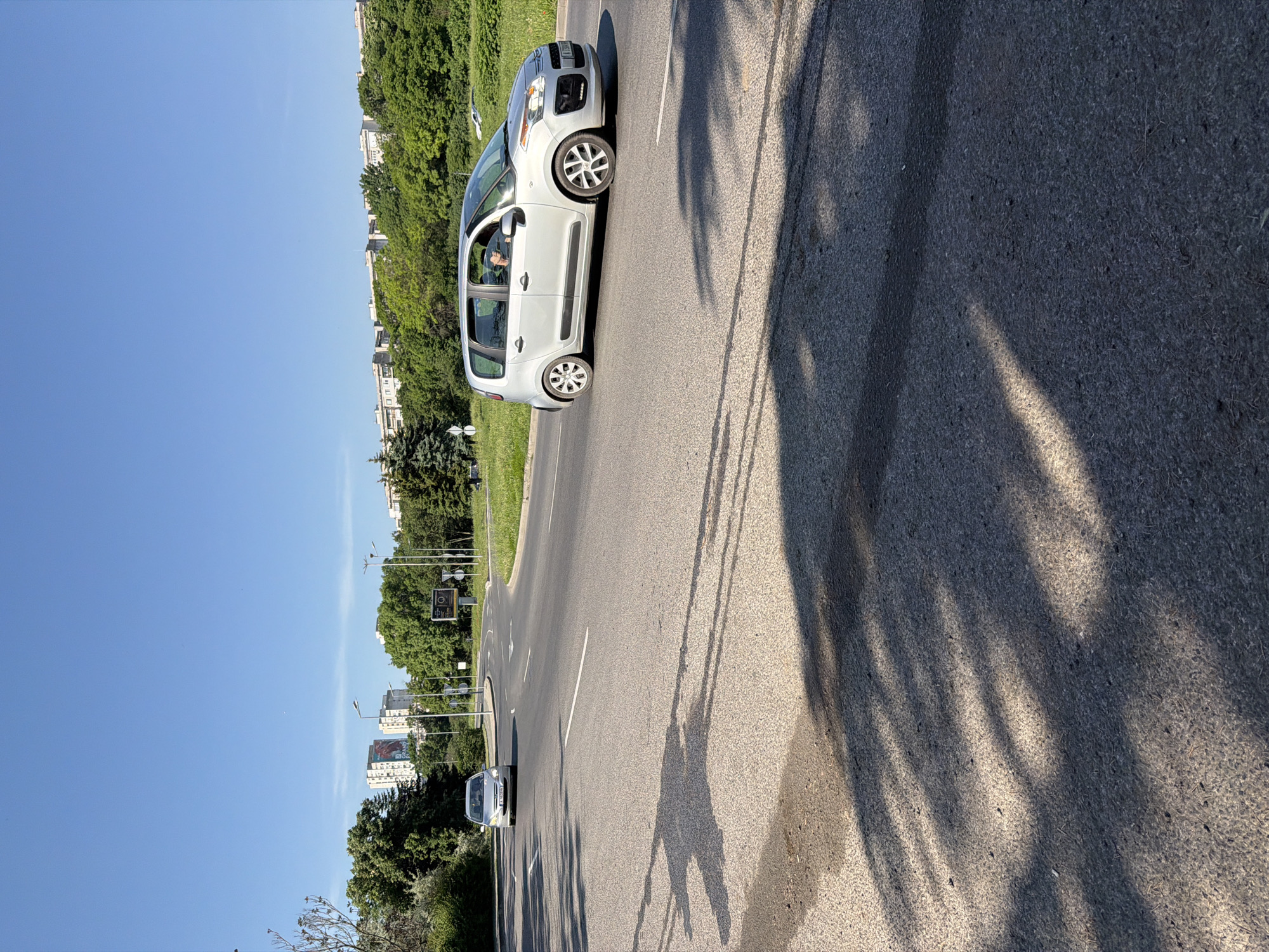 The image depicts a street scene in Burgas, Bulgaria. A light-colored Citroen C3 Picasso car is driving on the right side of a paved road. Another car is visible further down the road in the same direction. Trees line the roadside, casting shadows on the pavement. In the background, buildings are visible, and the sky is clear and blue. The presence of streetlights and signs indicates this is a typical urban roadway. The sun is shining brightly, evidenced by the distinct shadows. A person is visible in the driver seat of the Citroen. The road is marked with a white lane dividing line.