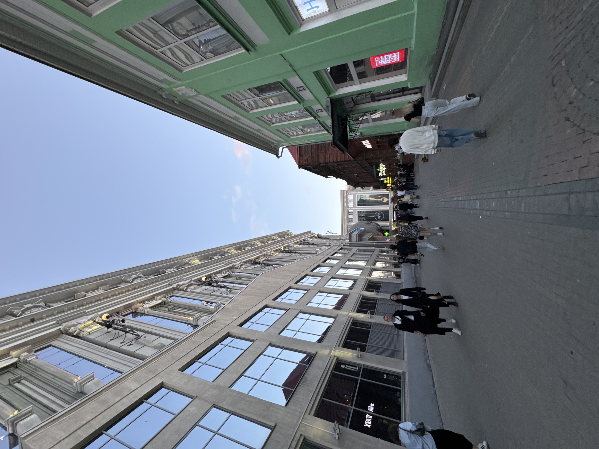 This is an image taken at street level looking upwards at buildings. The scene appears to be a city street with people walking by. The perspective is low and wide, with buildings on both the left and right sides of the frame.
On the left, a light-colored, ornate building with large windows dominates the lower part of the frame. These windows reflect the blue sky. The building has decorative architectural elements above the windows.
On the right, a light green building with a modern facade is visible. This building also has windows, and some signage is visible on it. Further down the street, more buildings and people can be seen.
There are several people walking on the sidewalk. They appear to be dressed in casual clothing. Some are walking in groups, while others are alone.
The time of day appears to be late afternoon or early evening, as the sky is a clear blue with a hint of pink near the horizon, suggesting a sunset. The weather appears to be pleasant.
Visible text includes a red sign with white lettering on the green building that says "OPENING HOURS". There are also signs with Cyrillic lettering visible further down the street, on other buildings, suggesting an urban setting in Russia. One sign on the light-colored building on the left has text in Cyrillic that reads "#HV ALIAX".
