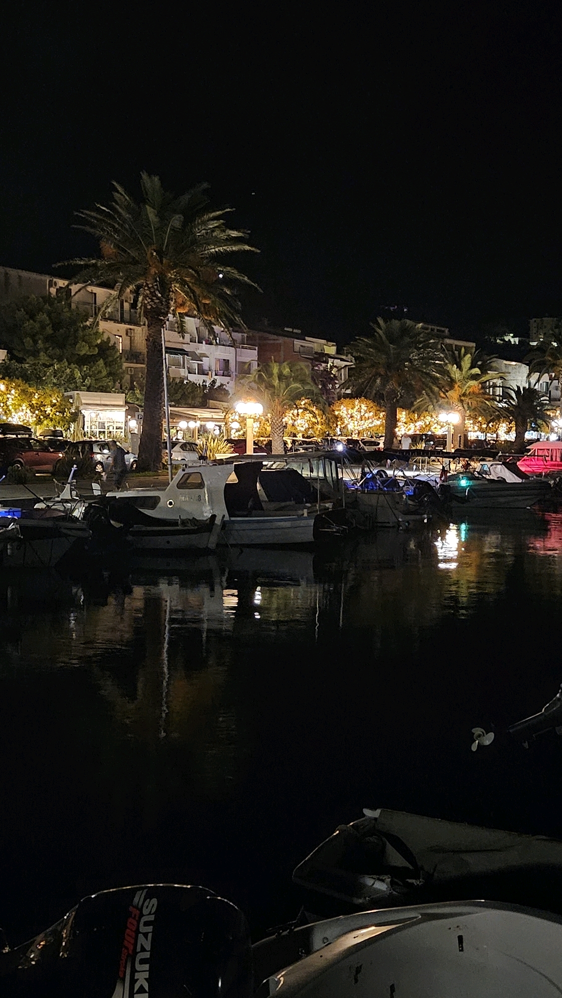 A nighttime scene captures a harbor in Općina Tučepi, Croatia. Several boats are moored, their forms silhouetted against the dark water. A prominent palm tree stands to the left, its fronds illuminated by ambient lights. Behind the harbor, buildings line the waterfront, with lights from windows and street lamps creating a warm glow amidst the darkness. The water reflects the lights from the buildings and the boats, creating a shimmering effect. In the foreground, the back of a boat with a "SUZUKI" outboard motor is visible.