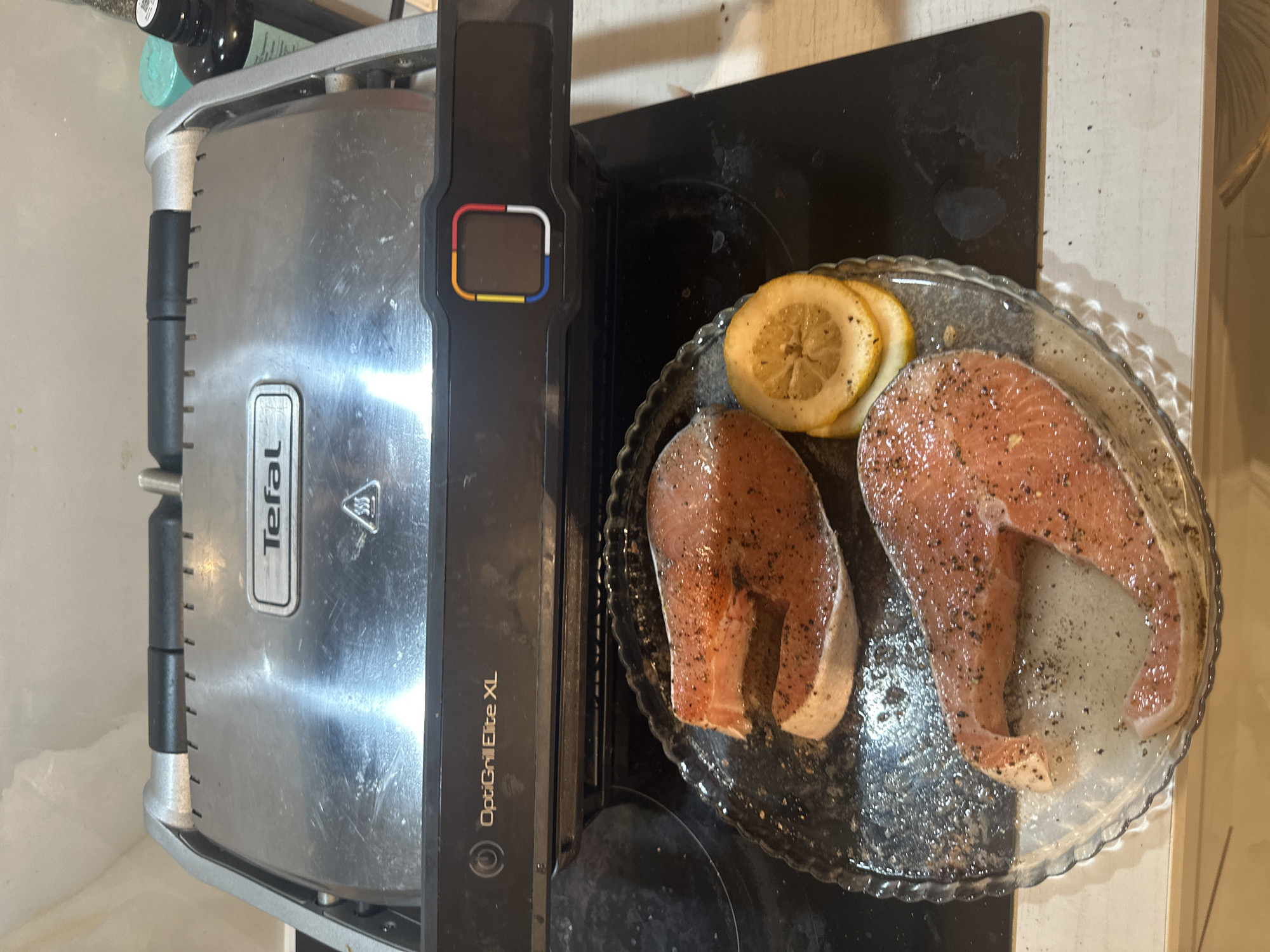 An overhead shot captures a kitchen scene featuring cooking preparation. On the left, a silver and black Tefal OptiGrill Elite XL electric grill is visible, its brushed stainless steel lid bearing the "Tefal" logo and a heat warning symbol. The side of the grill clearly displays "OptiGrill Elite XL." A distinctive control panel on the grill's handle features unlit colored segments – red, white, blue, and yellow – surrounding a central square, indicative of its smart grilling functions.
To the right of the grill, two raw, fresh salmon steaks are presented on a clear, scalloped glass plate. Both steaks are generously seasoned with black pepper and appear to be lightly oiled. Alongside the salmon are several fresh lemon slices, ready for use. The plate rests on a dark, potentially black, countertop that shows some minor splatters or smudges. The surrounding background includes light-colored wall or counter surfaces, placing the scene indoors, likely a home kitchen in Khimki, Russia. The image suggests a meal is actively being prepared.