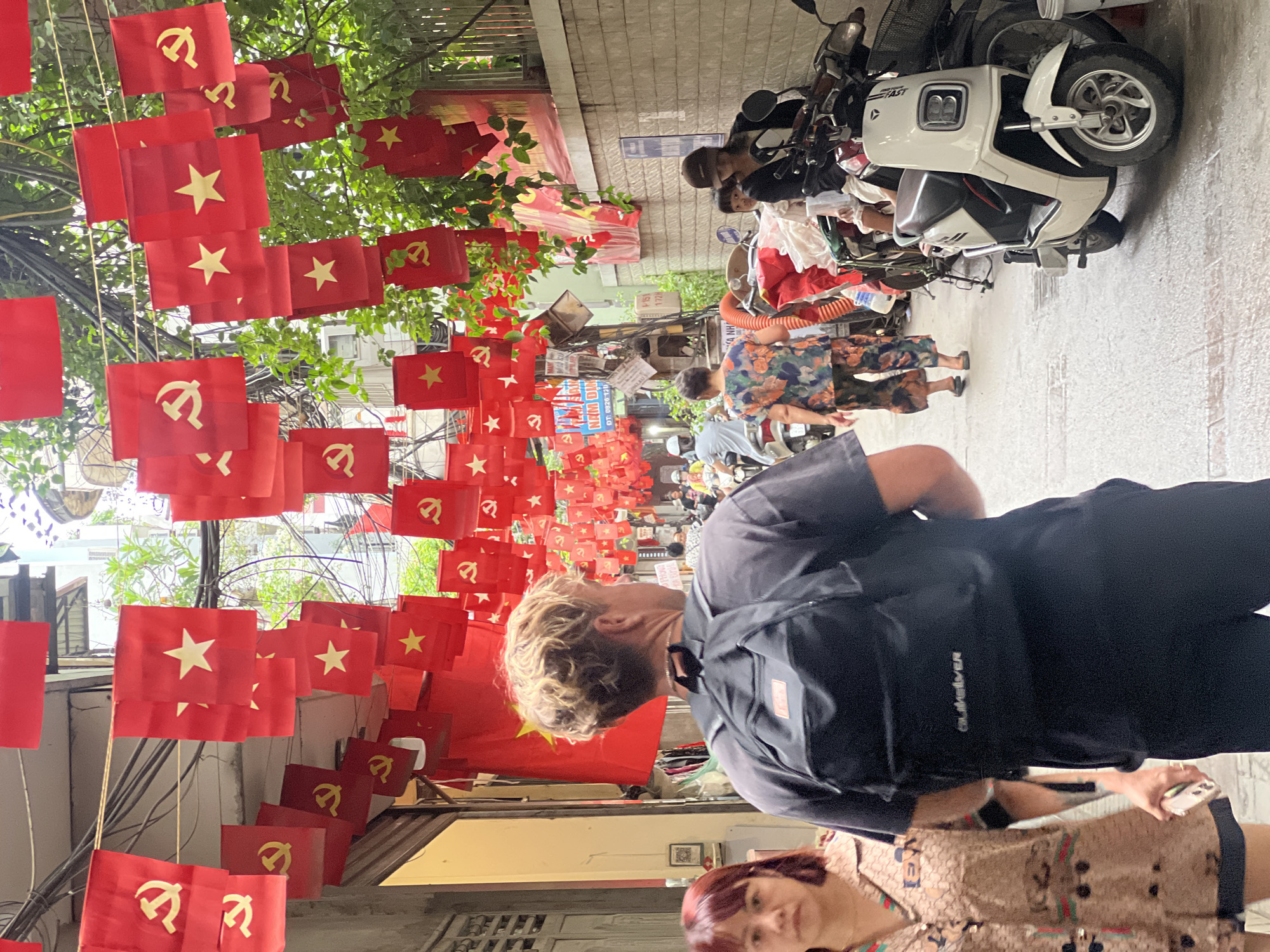 This image captures a street scene in Hanoi, Vietnam, adorned with numerous red flags featuring yellow stars and communist symbols, creating a festive or patriotic atmosphere. The flags are strung across a narrow alleyway, with some hanging from trees and overhead wires.
In the foreground, a man with blonde, curly hair wearing a black t-shirt and backpack walks away from the camera. To his left, a woman with short, dark red hair is partially visible, wearing a patterned shirt.
The alley is lined with buildings on either side. On the right, a brick wall is visible, and several motorbikes are parked along the side. One white motorbike is prominently featured in the mid-ground. People are seen walking and riding motorbikes further down the alley. The environment suggests a bustling urban setting, possibly a market or a residential area, with overhead wires and vegetation adding to the urban landscape. The overall impression is one of a vibrant, culturally rich, and busy street in Vietnam.