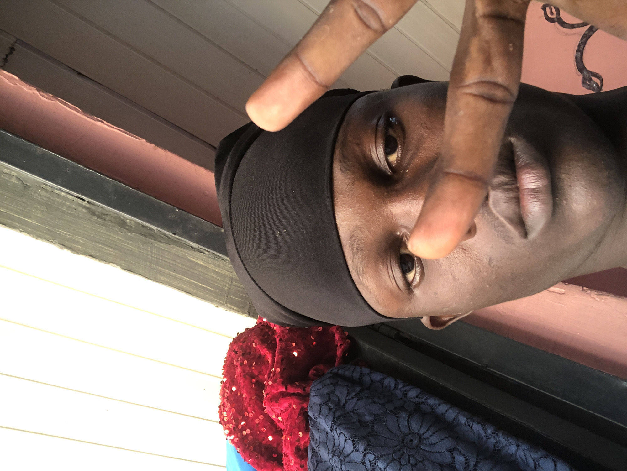 The image is a close-up, low-angle shot of a young Black person wearing a black durag. Their face is the primary subject, with their right eye visible and a finger held up in a peace sign gesture, partially obscuring their face. The person is looking towards the camera with a neutral expression.
In the background, there are horizontal wooden planks, suggesting an outdoor setting, possibly a porch or a wall. Below the person, a stack of fabric is visible, including a vibrant red sequined material and a dark blue lacy fabric.
The lighting appears natural, indicating daytime, with shadows suggesting it could be late afternoon. The overall impression is informal and personal, like a selfie. The context provided places the location in Ado, Nigeria. There is no visible text in the image.