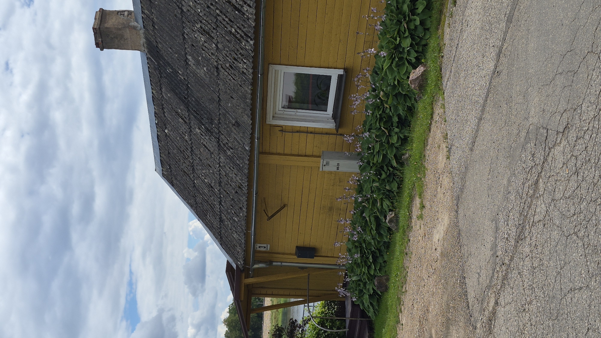 The image depicts a yellow wooden house with a corrugated roof and a brick chimney on the right side. A white-framed window is visible on the side of the house. In front of the house, there is a row of green plants with small purple flowers, bordered by rocks and grass. A gravel and asphalt driveway is in the foreground. The scene appears to be in a rural or suburban setting, with a body of water visible in the background to the left. The sky is cloudy. There are no people or animals present. Based on the architecture and the overall setting, this house could be located in Jēkabpils, Latvia, which is known for its historic wooden buildings and its location on the Daugava River.