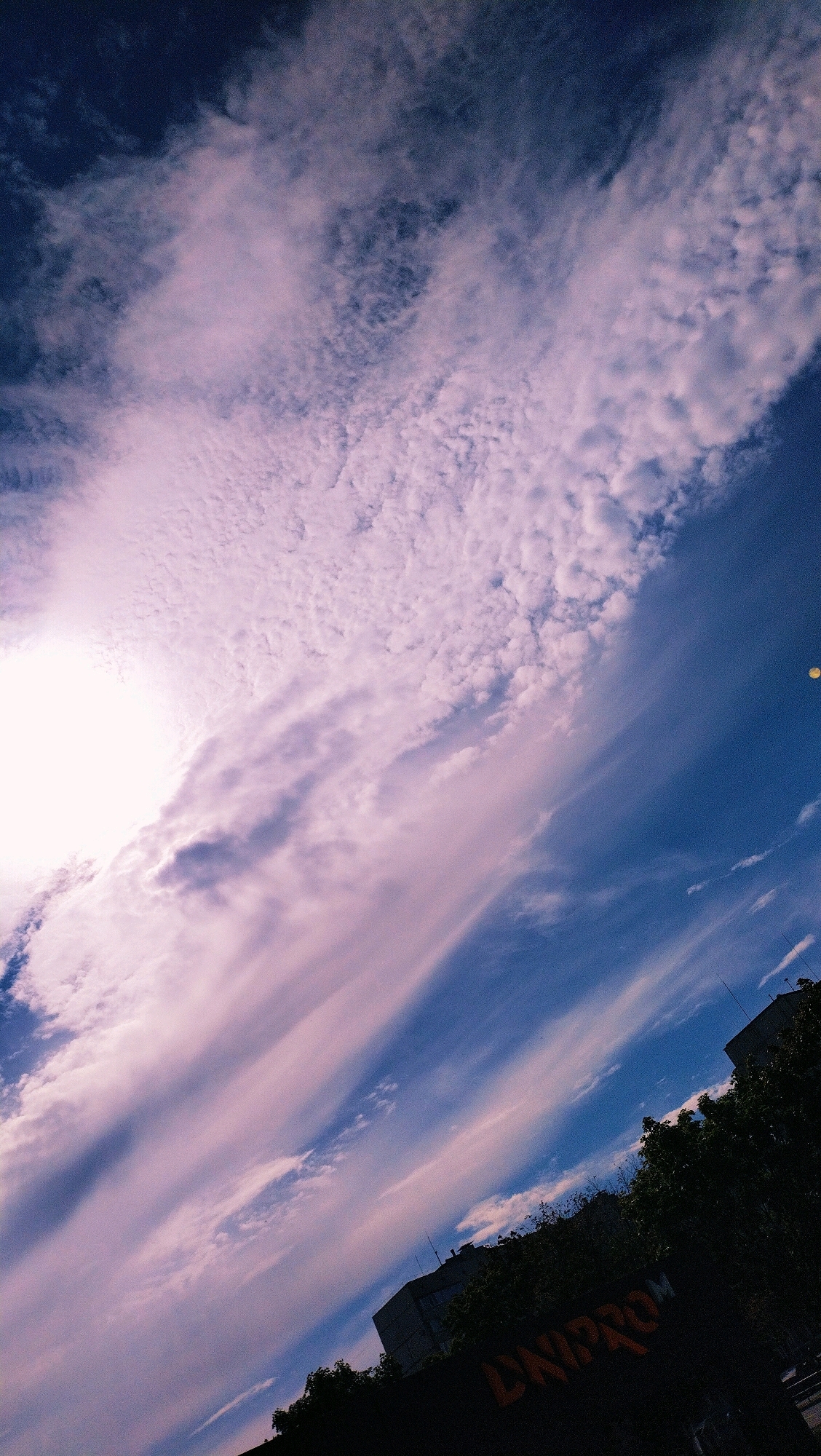 The image captures a tilted view of a sky with clouds and buildings in the foreground, possibly in Horishni Plavni, Ukraine. The sky is a vibrant blue, with wispy, pink-tinged clouds scattered across it. The sun is partially visible in the upper left corner, casting a bright glow. In the lower portion of the image, dark silhouettes of trees and buildings are visible against the bright sky. One building has orange graffiti on it that reads "DNIPRO". The overall impression is a dynamic and dramatic sky scene over an urban landscape.
