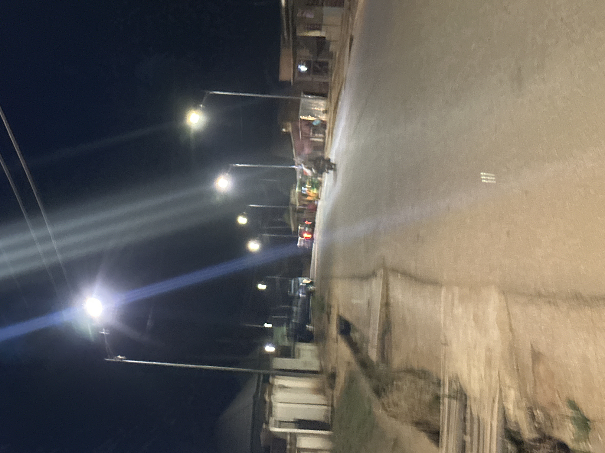 The image shows a street scene at night in Bukuru, Nigeria. Streetlights illuminate the road and the buildings lining it. A motorcycle is visible on the road, and there are parked vehicles further down. The sidewalk is present on the side closest to the viewer. Buildings with lights visible in the windows are on the opposite side of the road. Power lines are visible above. The scene is blurry.