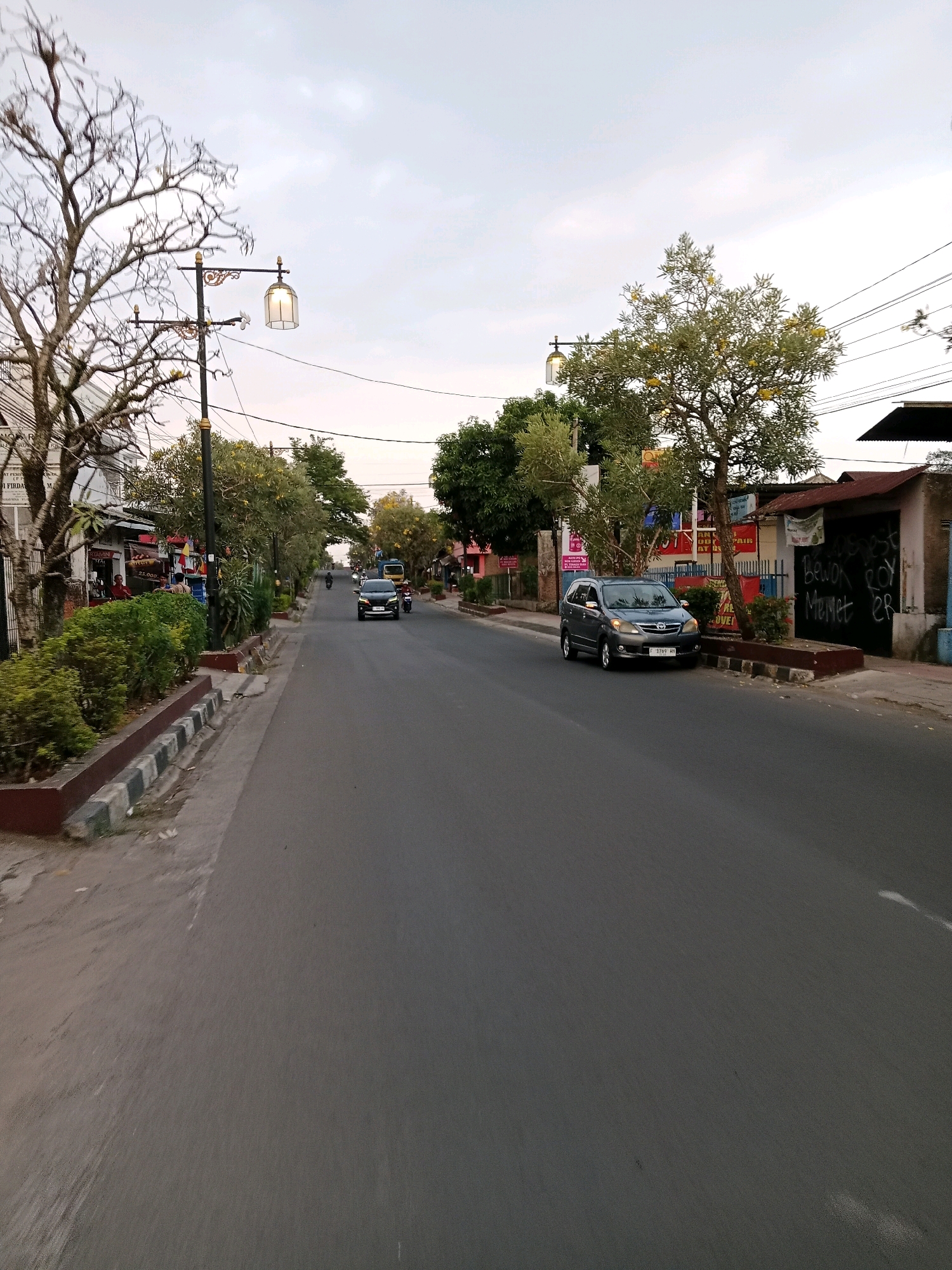The image shows a street scene in the city of Cianjur, Indonesia, on what appears to be an overcast day. The perspective is from a moving vehicle, looking down a paved road lined with trees and buildings.
On the left side of the road, there is a sidewalk area with bushes and some low-lying trees. A few shops or stalls are visible behind the foliage. A distinctive ornate street lamp with a caged light fixture stands on the sidewalk. Further down the street, more trees and buildings line the road.
On the right side of the road, there are more trees, some of which have yellow flowers. A dark gray car is parked on the side of the road, and its license plate reads "F 1769 UH". Next to the parked car is a building with a corrugated metal roof and a large black gate with graffiti. The graffiti includes the words "Bewok ROY Memet ER".
Traffic on the road includes the parked car, a dark-colored car in motion further down the street, a yellow vehicle that appears to be a small bus or van, a motorcycle with a rider, and possibly other vehicles in the distance. The sky is cloudy and light gray. The overall impression is that of a typical urban street in Indonesia, likely during daytime.