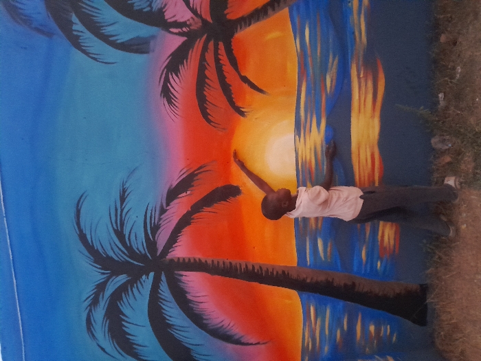 In Kirana, Nigeria, a young male individual stands outdoors before a vibrant mural painted on a wall. The mural depicts a tropical sunset scene with a gradient sky transitioning from blue at the top to orange and red at the horizon, featuring a prominent setting sun on the right side. Silhouetted black palm trees are painted on the left and right edges of the mural. Below the horizon, the mural shows ocean water with blue, gold, and white reflections. The child, wearing a light-colored short-sleeved collared shirt and dark trousers, is positioned in the lower-center, facing right. His right arm is extended upwards, hand reaching towards the painted sun, while his left arm is bent forward. He stands on a patch of dry grass and dirt, with some scattered debris visible on the ground to his right.