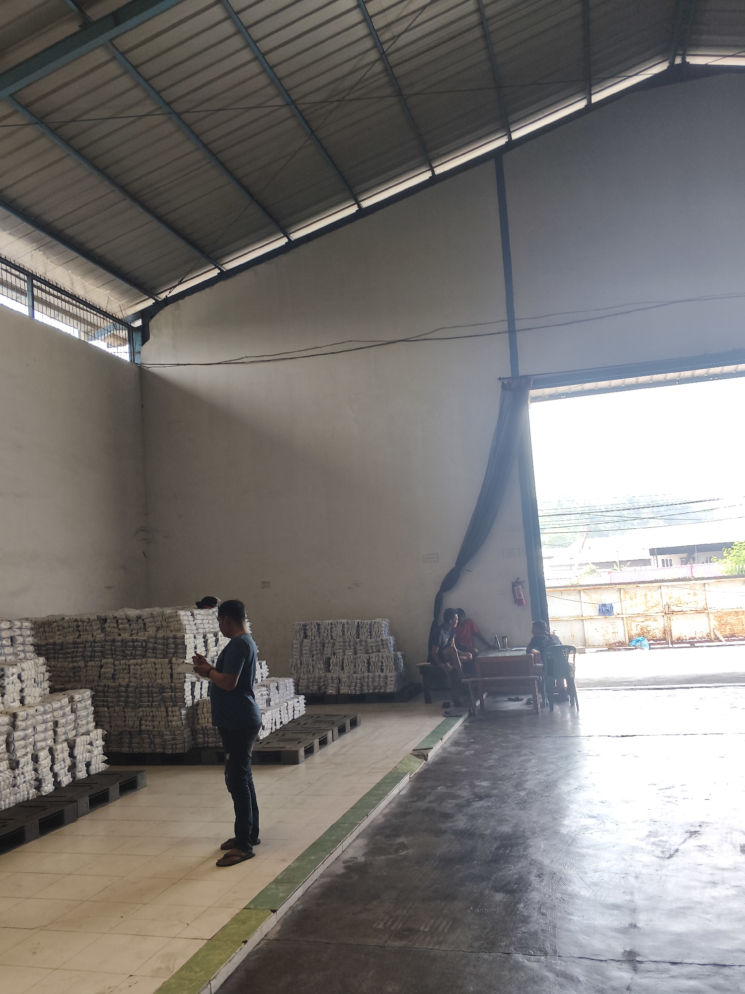 This is a full shot, eye-level, outdoor photograph of the interior of a warehouse with an open loading dock in Bandar Lampung, Indonesia. The warehouse has a metal roof with visible beams and supports. Stacks of white boxes are piled high on pallets. A man stands in the foreground, looking at his phone. In the background, several other people are seated around a table near the loading dock. The loading dock opens to a bright, outdoor scene with some buildings and vegetation visible. A fire extinguisher is mounted on the wall to the left of the loading dock. The floor is a combination of tiled and concrete surfaces. The lighting is bright, with sunlight streaming in from the open loading dock.
