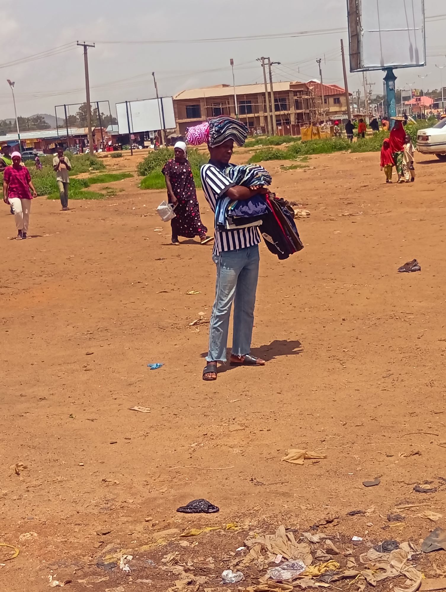 In Bukuru, Nigeria, a marketplace scene unfolds on a dusty, uneven dirt ground. A young man, dressed in a striped shirt and blue jeans, stands prominently in the foreground, balancing a stack of folded fabrics on his head and carrying a bag of clothes in his right arm. Behind him, several other individuals are visible. To his left, a woman in a red top and white trousers walks away from the camera. Further in the background, a woman in a dark patterned dress carries a white bag, and two young children in red and beige attire walk alongside a woman in a red headscarf. The backdrop features a cluster of buildings, some under construction, and overhead utility lines. The environment is littered with various debris, including plastic bags, papers, and other discarded items, indicative of a busy, potentially informal, commercial area.