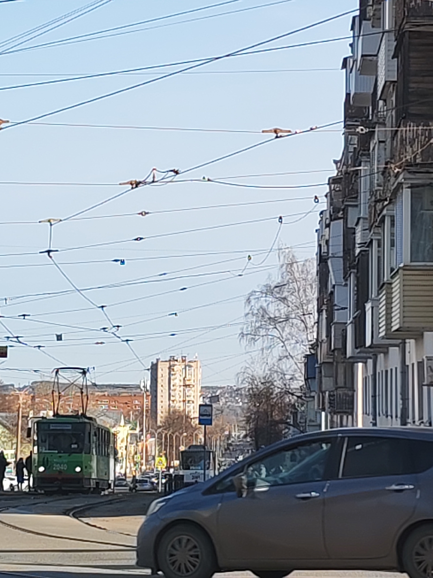 The image depicts a street scene in Izhevsk, Russia. A green tram, numbered 2040, is on the tracks, approaching the viewer. A blue Nissan hatchback is in the foreground, slightly obscuring the view. In the background is a tall apartment building. Overhead, the complex network of tram wires dominates the sky. Other vehicles are visible on the street. People are walking near the tram stop. The day appears sunny.
