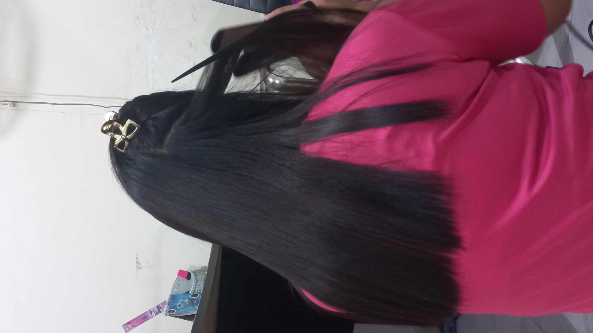 The image, taken indoors in what appears to be a home or personal grooming space in Jetis Indah, Indonesia, shows the back of a person with long, straight, dark hair. The hair is neatly sectioned, with a golden bow-shaped hair clip securing a portion at the top. A thin comb is being used to style another section of hair. The person is wearing a bright pink t-shirt. In the background, a white wall is visible, along with a shelf containing some stationery items, including notebooks and a ruler. The overall scene suggests a hair styling activity in progress.