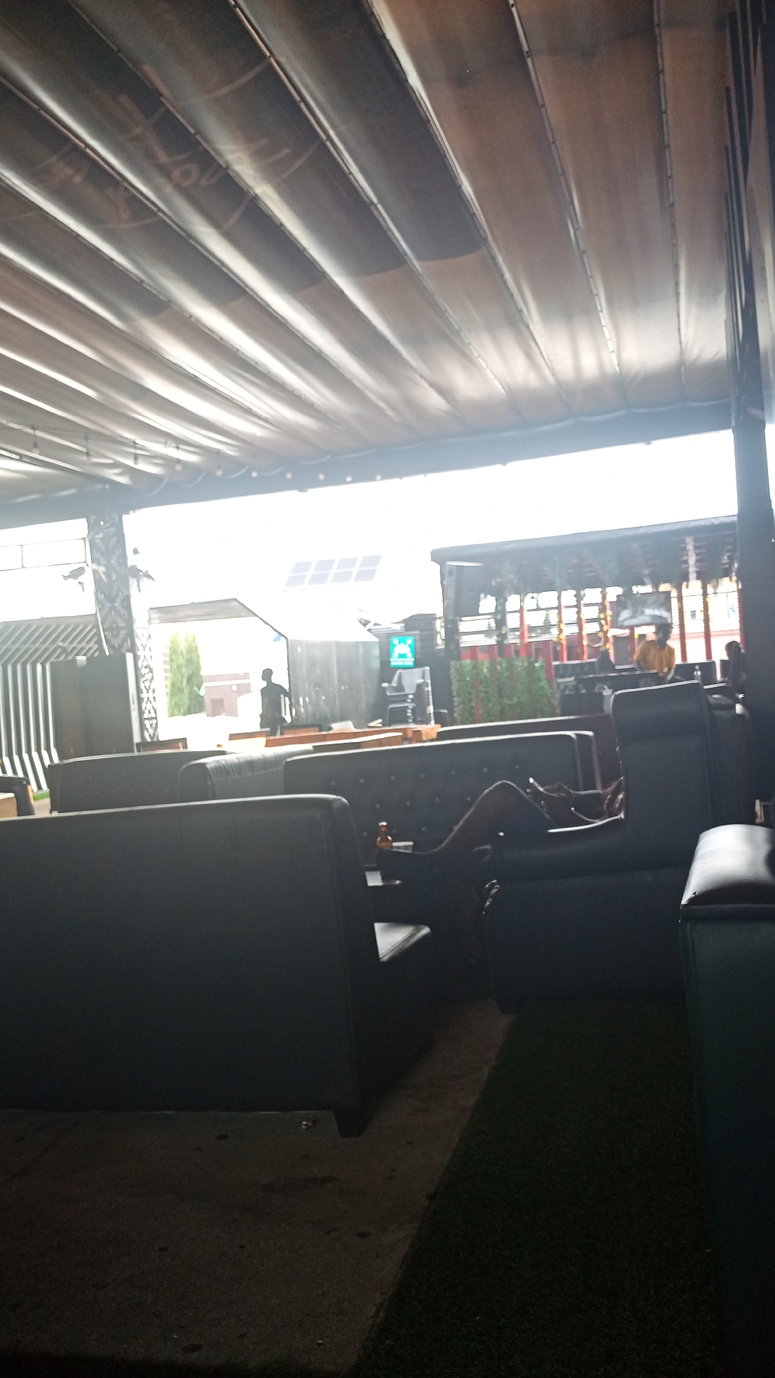 This media file depicts an outdoor lounge area, possibly at a venue in Howayi, Nigeria. The scene is illuminated by natural daylight, suggesting it is daytime. The area features dark, plush seating, including sofas and armchairs, arranged around tables. The ceiling is covered by a retractable awning made of corrugated material, which casts patterned shadows across the space.
In the background, there is a structure with solar panels on its roof, a stage or platform area, and what appears to be a DJ booth. Several people are visible. One individual is seen lounging on a sofa, appearing relaxed. Other figures are further in the background, engaged in various activities, some of whom appear to be staff or performers. The atmosphere seems casual and social, likely a place for entertainment or relaxation. The presence of artificial turf on the ground suggests an outdoor recreational or hospitality setting. There is no visible text in the image.