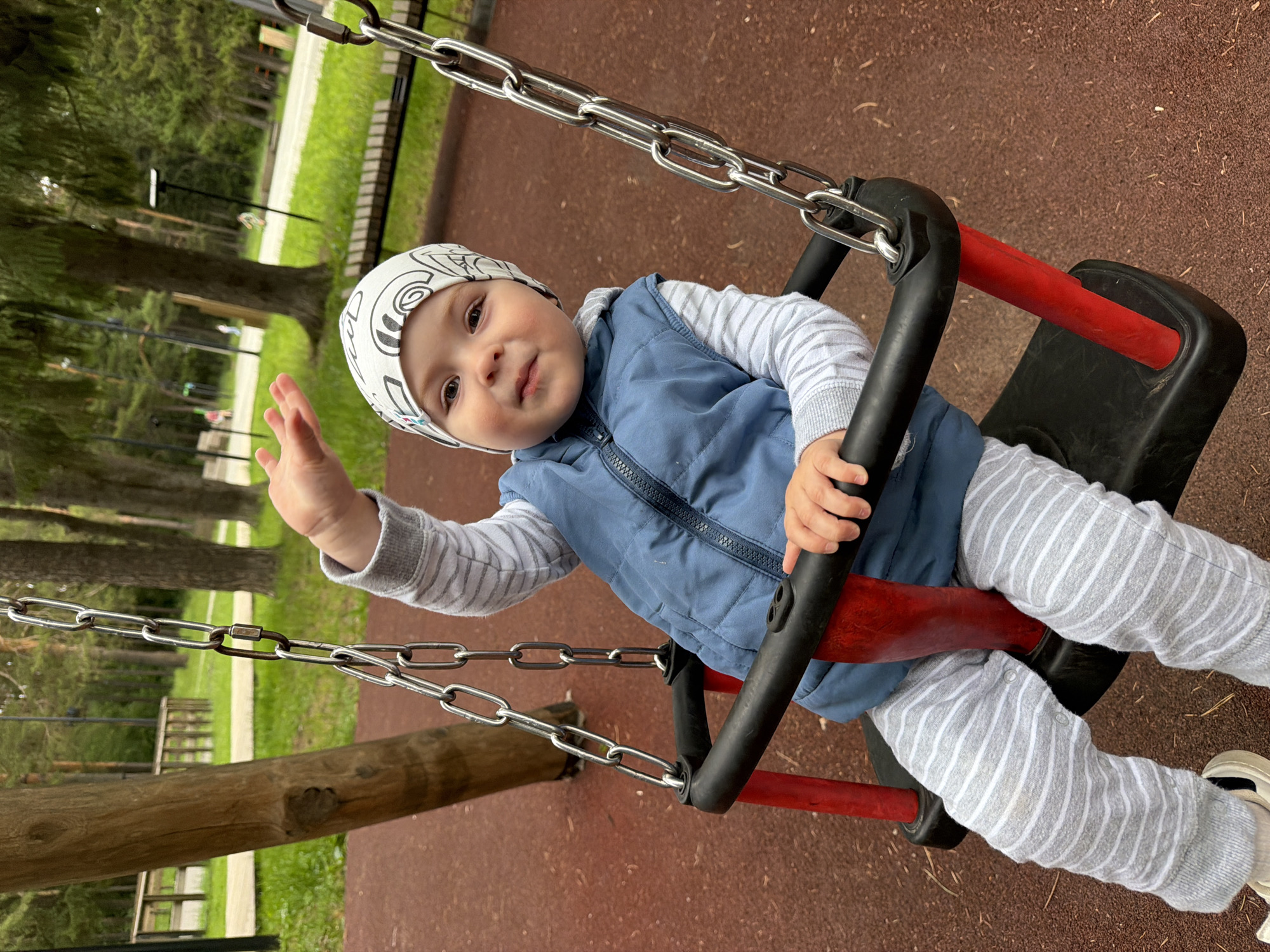 A close-up shot captures a baby sitting on a playground swing. The baby is wearing a blue vest over a striped long-sleeved shirt and striped pants, along with a white hat adorned with a black car pattern. The baby's right arm is raised in a waving gesture, and their left hand rests on the swing's bar. The swing itself has a black seat with red supports and is suspended by metal chains. The background reveals a park-like setting with trees and a path, consistent with an outdoor playground area. The ground beneath the swing is covered in red rubber mulch. The scene is set in Votkinsk, Russia.