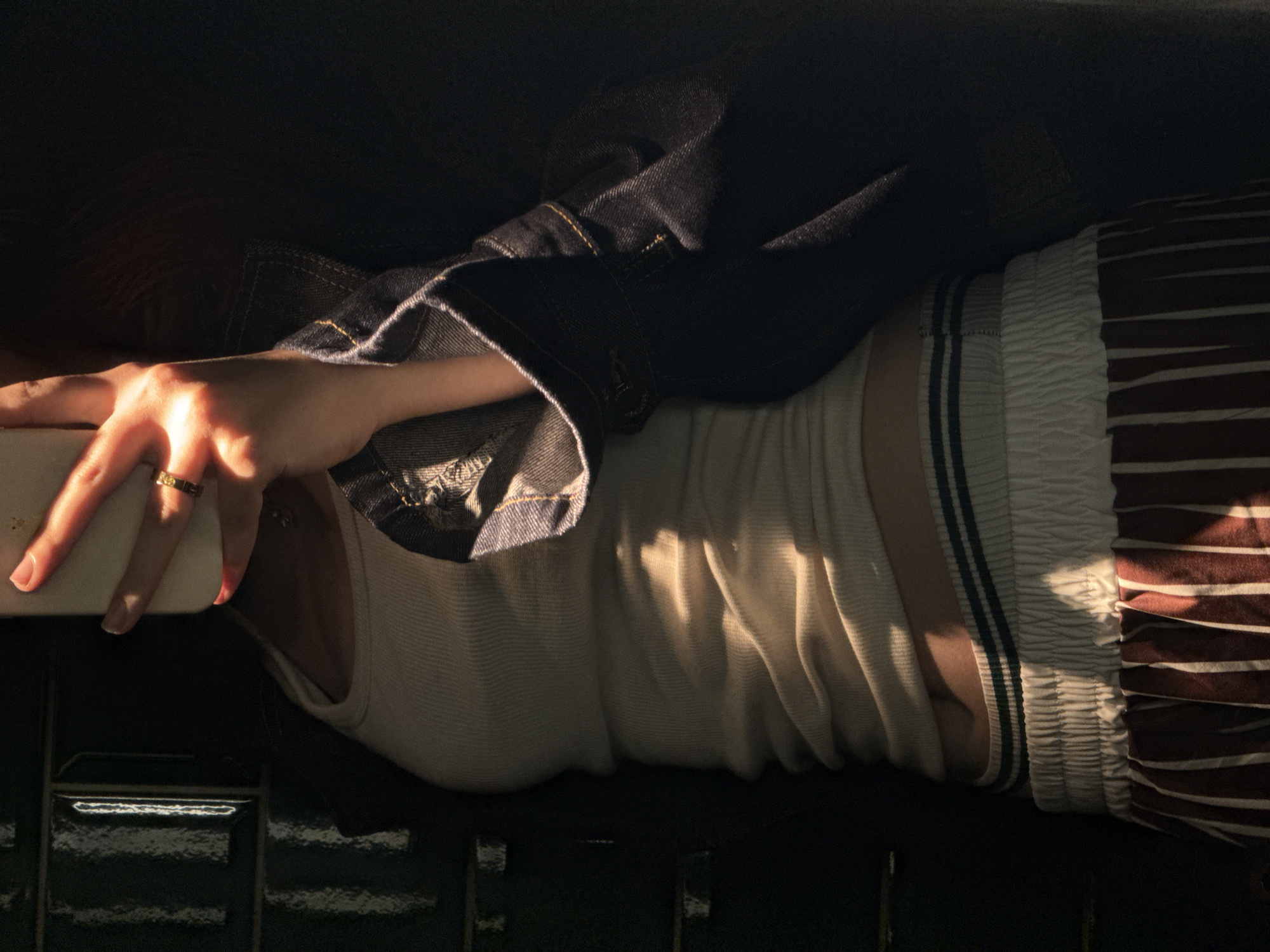A person is lying down, wearing a white tank top, a dark denim jacket, and striped pants. The person is holding a phone in their hand. The scene is partially illuminated by sunlight. The location in Yerevan, Armenia, is not evident from the image.