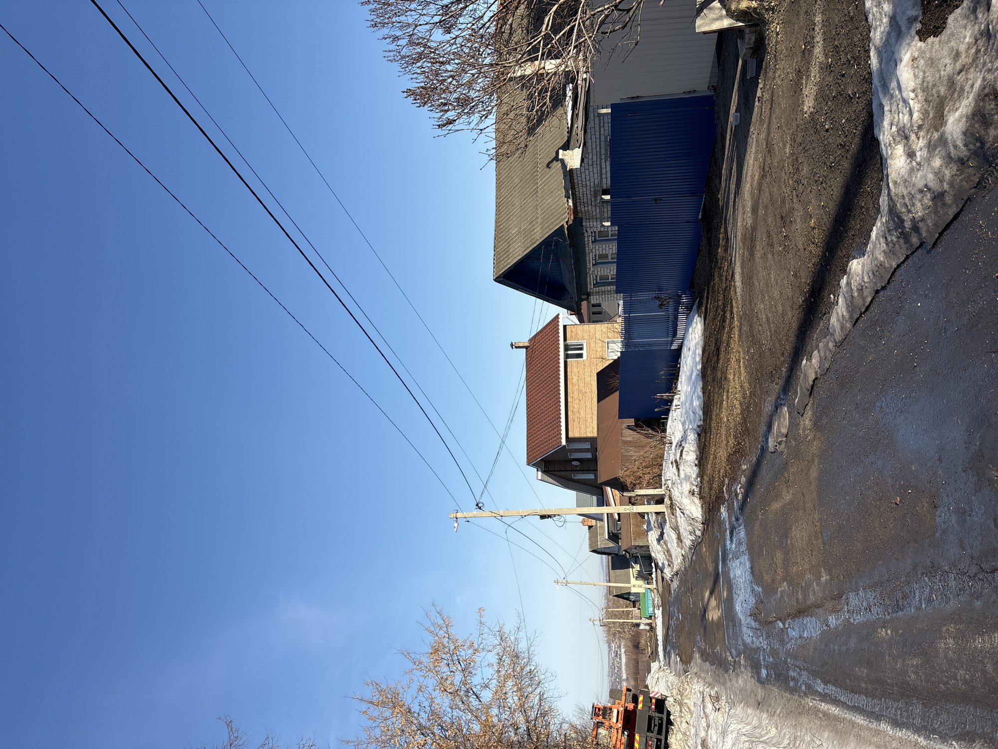 The image displays a residential street in Ulyanovsk, Russia, under a clear, bright blue sky. The unpaved road, viewed from a slightly elevated position, is muddy and uneven with prominent patches of melting snow and ice along its edges and in depressions.
On the left side of the frame, the rear portion of an orange heavy-duty truck, possibly a flatbed or construction vehicle, is partially visible, parked on a snow-covered verge. Further down this side, dormant deciduous trees with some clinging dry leaves line the road, and several single-story and two-story houses with varied roofing (e.g., green, grey) recede into the distance.
On the right side, a dark blue corrugated metal fence separates the properties from the street. Behind this fence, a white brick house with blue trim and a dark grey corrugated roof is visible in the midground, adjacent to a more modern two-story brick house featuring a brown tiled roof. Utility poles, some concrete and some wooden, line both sides of the street, supporting multiple black power lines that traverse the upper portion of the frame. A street light fixture is mounted on a concrete pole on the right. No individuals, animals, or direct actions are depicted. The scene suggests a quiet, suburban environment transitioning from winter to spring.