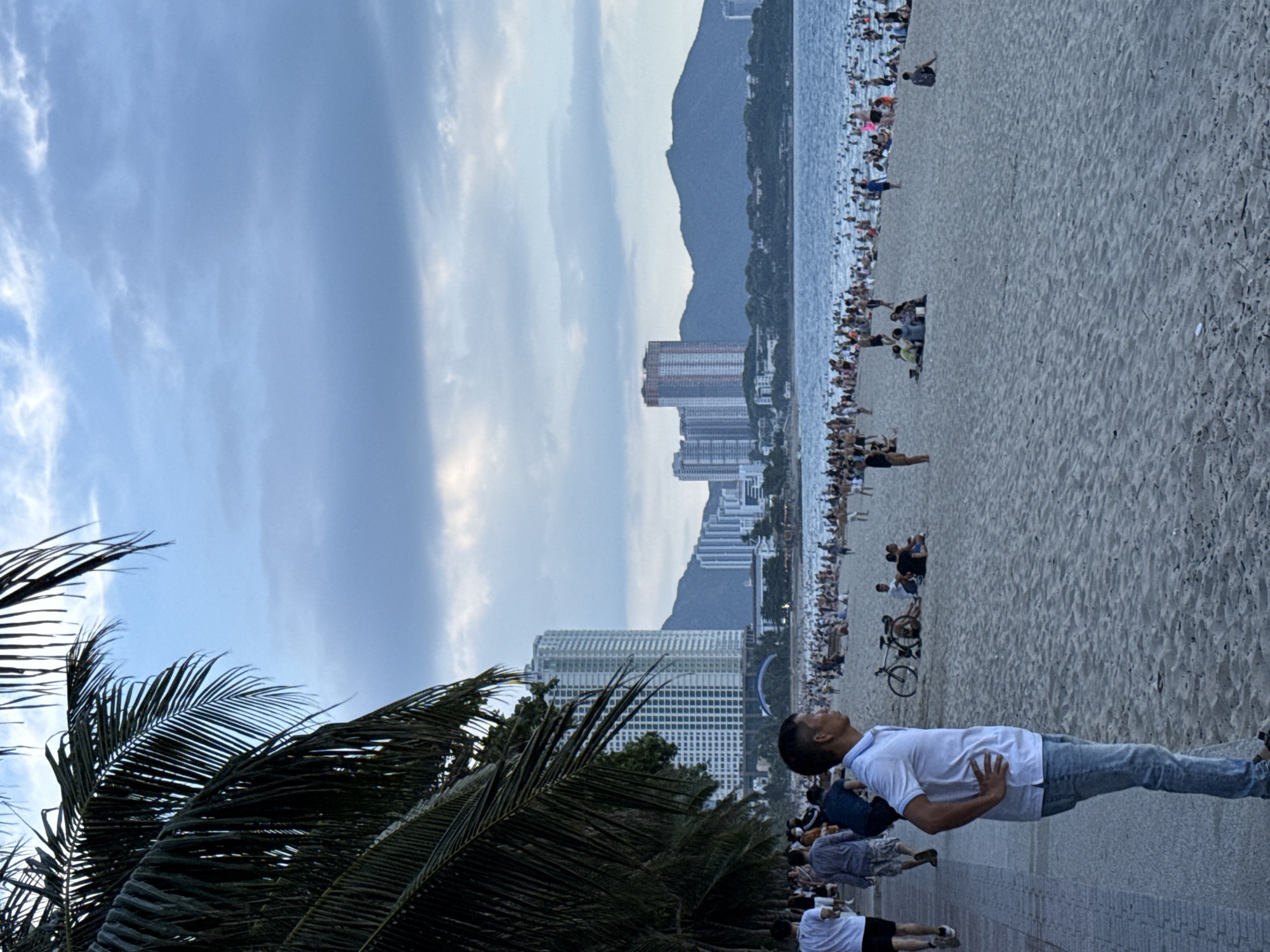 This is a photograph of a beach in Nha Trang Ward, Vietnam, taken during the late afternoon or early evening. The scene is populated by a large number of people enjoying the beach and the water. In the foreground, a man in a white polo shirt and blue jeans stands with his hands on his hips, looking out towards the ocean. Behind him, the sandy beach is filled with beachgoers, some sitting in groups, others walking or playing. A bicycle is parked on the sand. The ocean is calm, with many people swimming or wading. In the background, a line of tall buildings, including high-rise apartments and hotels, are visible along the coastline, with mountains rising behind them. Palm trees line the walkway to the left of the frame, partially obscuring the view of the buildings. The sky is a mix of blue and grey clouds, suggesting the sun may be setting or has recently set.