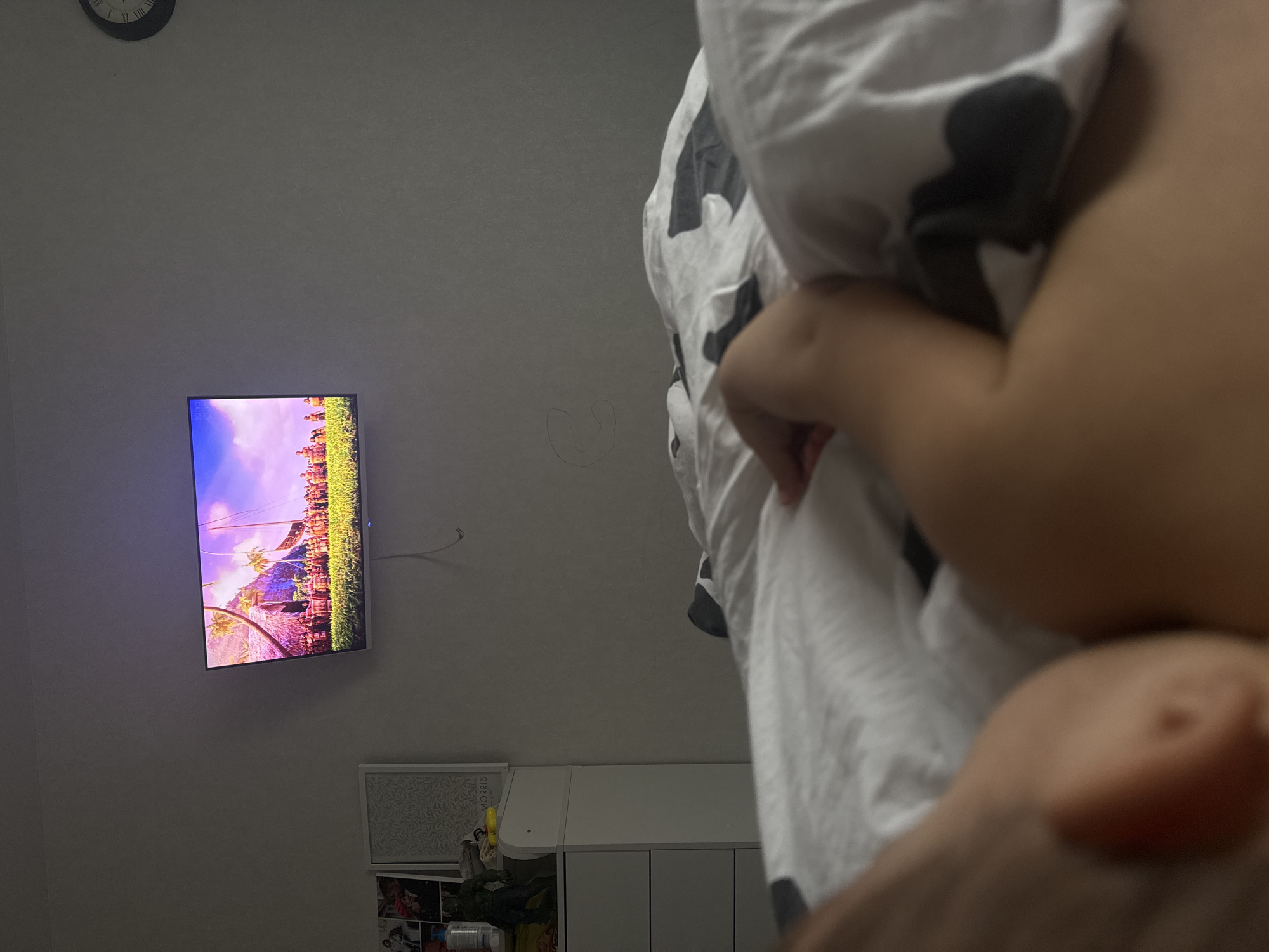 The image shows a person in bed with a white duvet cover patterned with black spots. A television mounted on the wall is displaying a colorful scene, potentially a landscape. Below the television, there is a piece of art. The top left corner of the image shows an analogue clock. There are no identifiable details to confirm the specific location within Chaikovsky, Russia.