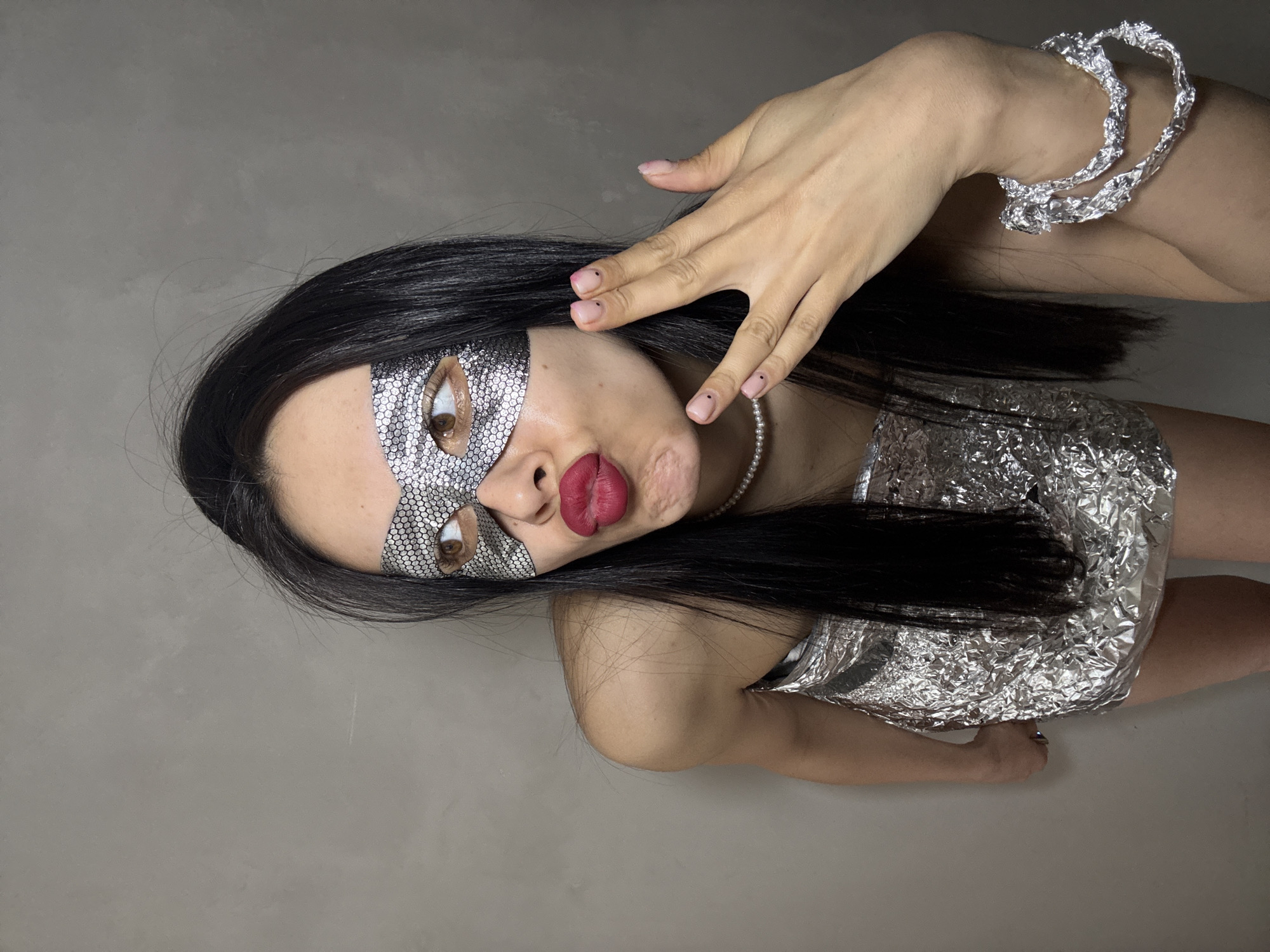 A woman with long dark hair, wearing a silver sequined mask over her eyes and a silver foil top, poses against a plain grey background. She is also wearing a pearl necklace and a silver foil bracelet. Her right hand is raised near her face. Her lips are pursed. The location is not discernible from the image and cannot be identified as Votkinsk, Russia.