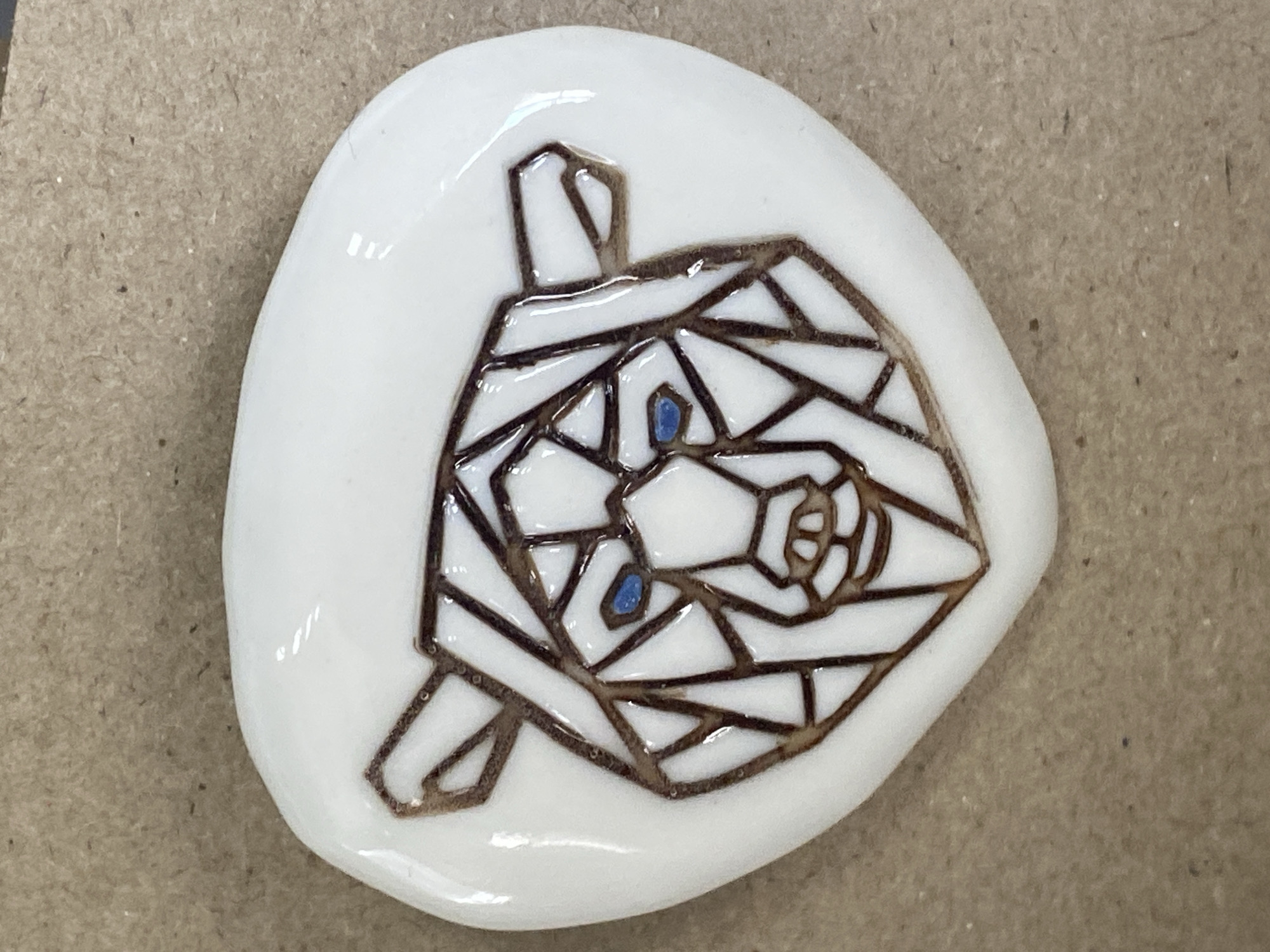 A white, irregular-shaped stone has a geometric design etched onto its surface. The design is a stylized depiction of a dog's head. The lines forming the geometric shapes are brown, and the dog's eyes are colored blue. The stone sits on a tan or brown surface. The location is unknown, but based on the prompt it may be Yekaterinburg, Russia.
