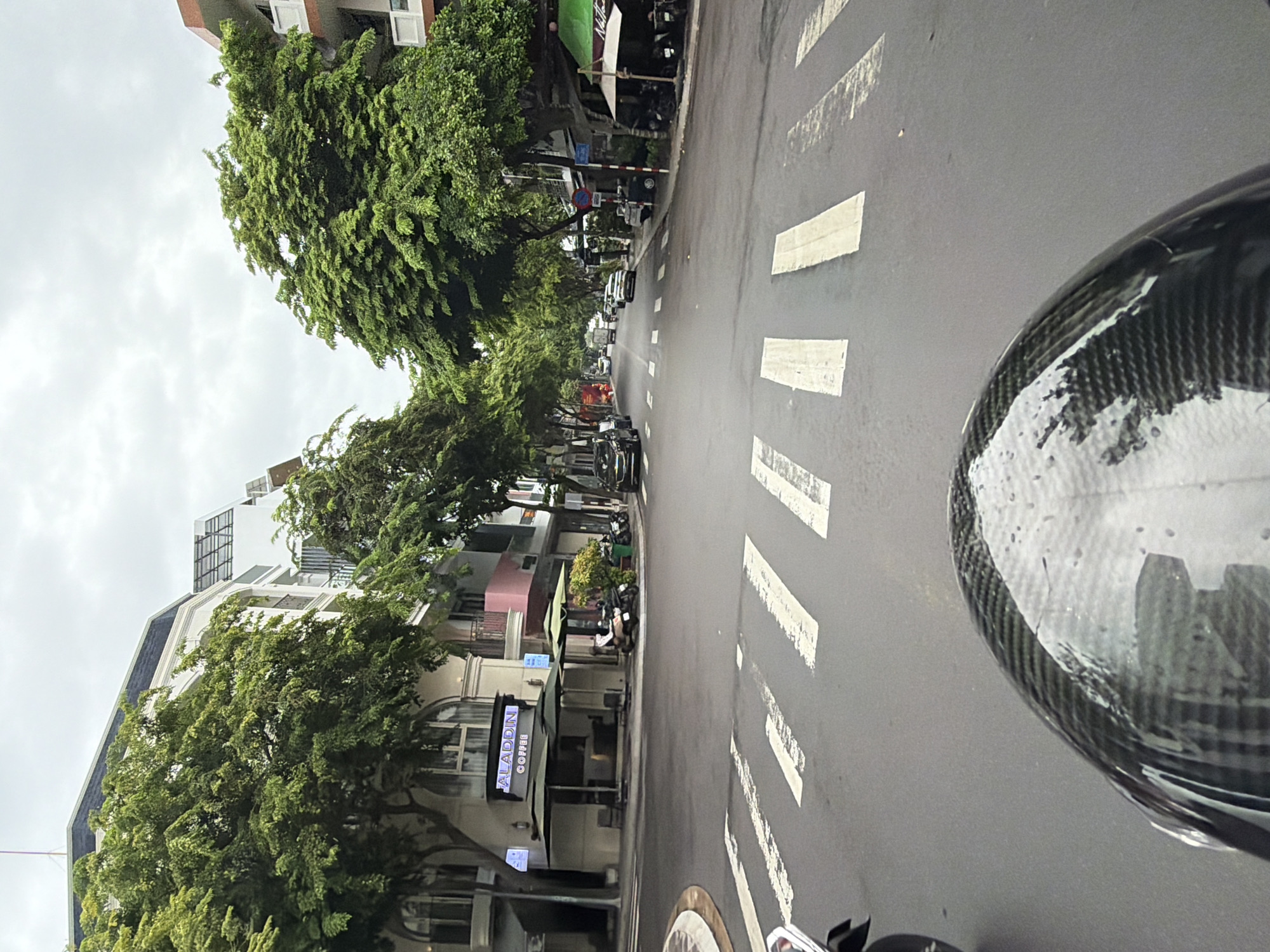 The image captures a street scene in Nha Trang, Vietnam, viewed from the perspective of someone riding a motorcycle. The foreground is dominated by the curved, carbon fiber-patterned surface of a motorcycle helmet, reflecting the sky and surrounding buildings. A pedestrian crossing, marked by thick white stripes, is visible on the asphalt road.
On the left side of the street, a building with a light-colored facade features arched windows and a sign reading "ALADDIN COFFEE" above a storefront with outdoor seating and umbrellas. Several motorcycles are parked nearby. Mature trees with lush green foliage line the street, casting shadows.
The road extends into the distance, with a moderate amount of traffic including cars and motorcycles. Buildings of varying heights and architectural styles are situated along both sides of the street. The sky is overcast, suggesting a cloudy day. The overall impression is of a typical urban environment in Vietnam.