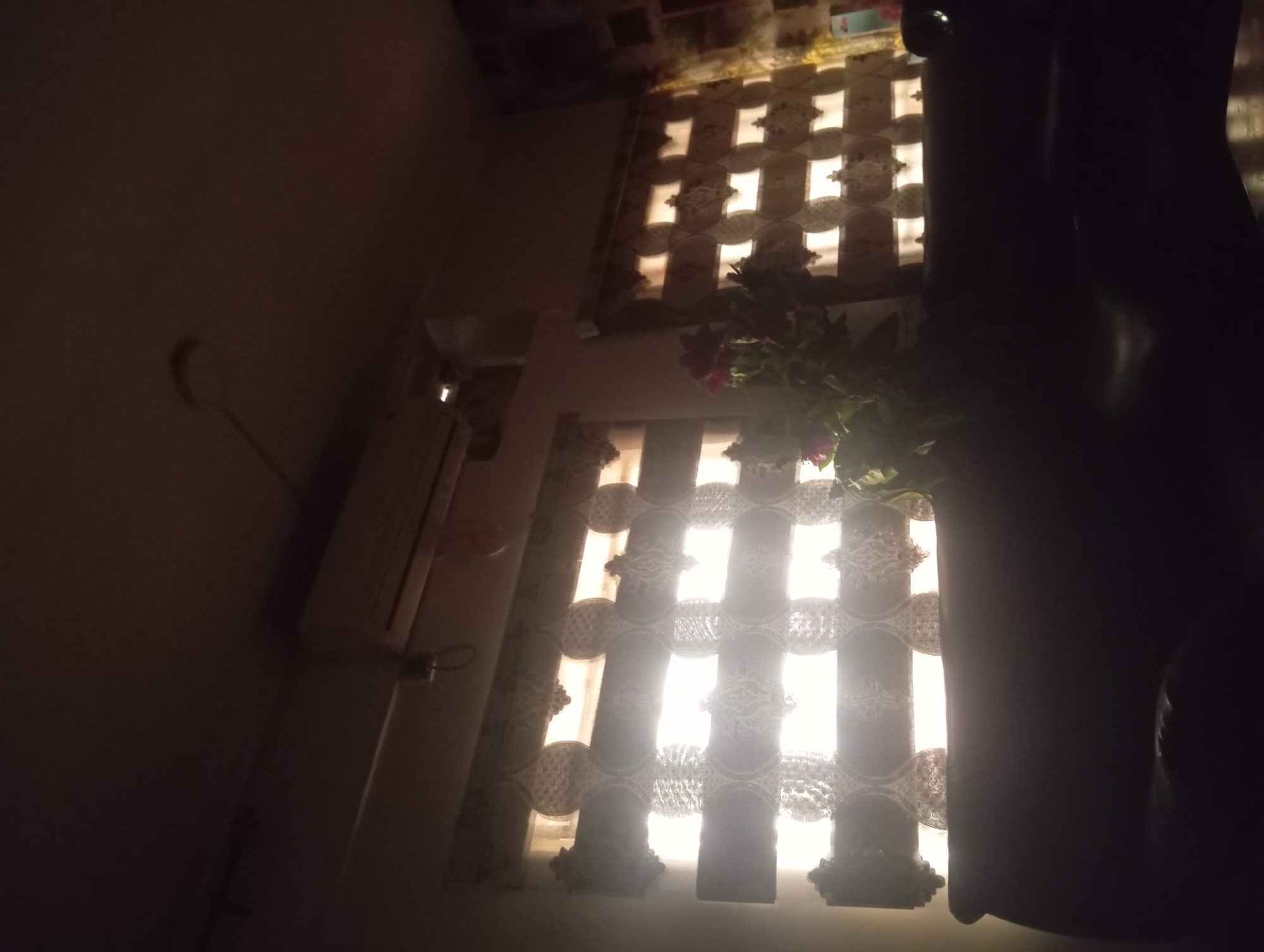 The image depicts an indoor scene, likely a room, with natural light filtering through two windows. The perspective is from a low angle, looking upwards and to the side.
The dominant features are the windows, covered by decorative curtains that create a patterned effect with the light. The curtains have a lace-like quality, with circular and cross-like motifs that allow bright light to pass through, casting distinct shapes on the wall and floor. One window is partially obscured by a dark, curtain-like object on the right side of the frame, which appears to be a thick curtain or possibly a piece of furniture covered in fabric.
In the foreground, on the left side of the image, a portion of a light-colored object, possibly an air conditioner unit, is visible. It is mounted high on the wall and has a rectangular shape. There is also a hint of a metallic handle or lever attached to it. Further to the left, a circular, pale object is seen on the wall, its purpose unclear.
A cluster of plants with dark pink or magenta flowers is positioned near the top right window, their leaves and blooms partially illuminated by the light.
The overall lighting suggests it is either early morning or late afternoon, with sunlight creating a warm glow. The scene appears to be quiet and still, with no visible people or active events. The emotions conveyed are tranquil and perhaps a bit mysterious due to the interplay of light and shadow. There is no visible text in the image.
