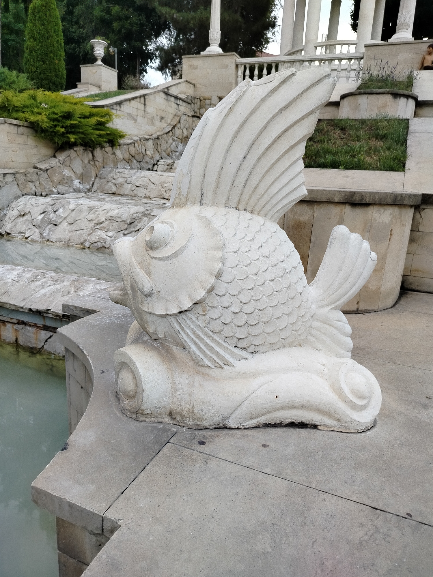The image shows a white concrete sculpture of a stylized fish resting on a tiered concrete structure adjacent to a body of water. The fish sculpture is detailed with scales and fins. In the background, there are stone stairs, lush green foliage, and classical architectural elements including columns and balustrades, suggesting an outdoor park or garden setting. There is a person partially visible in the upper right background. The location is the Stephen the Great Central Park in Chișinău, Moldova, specifically near the cascade fountains.