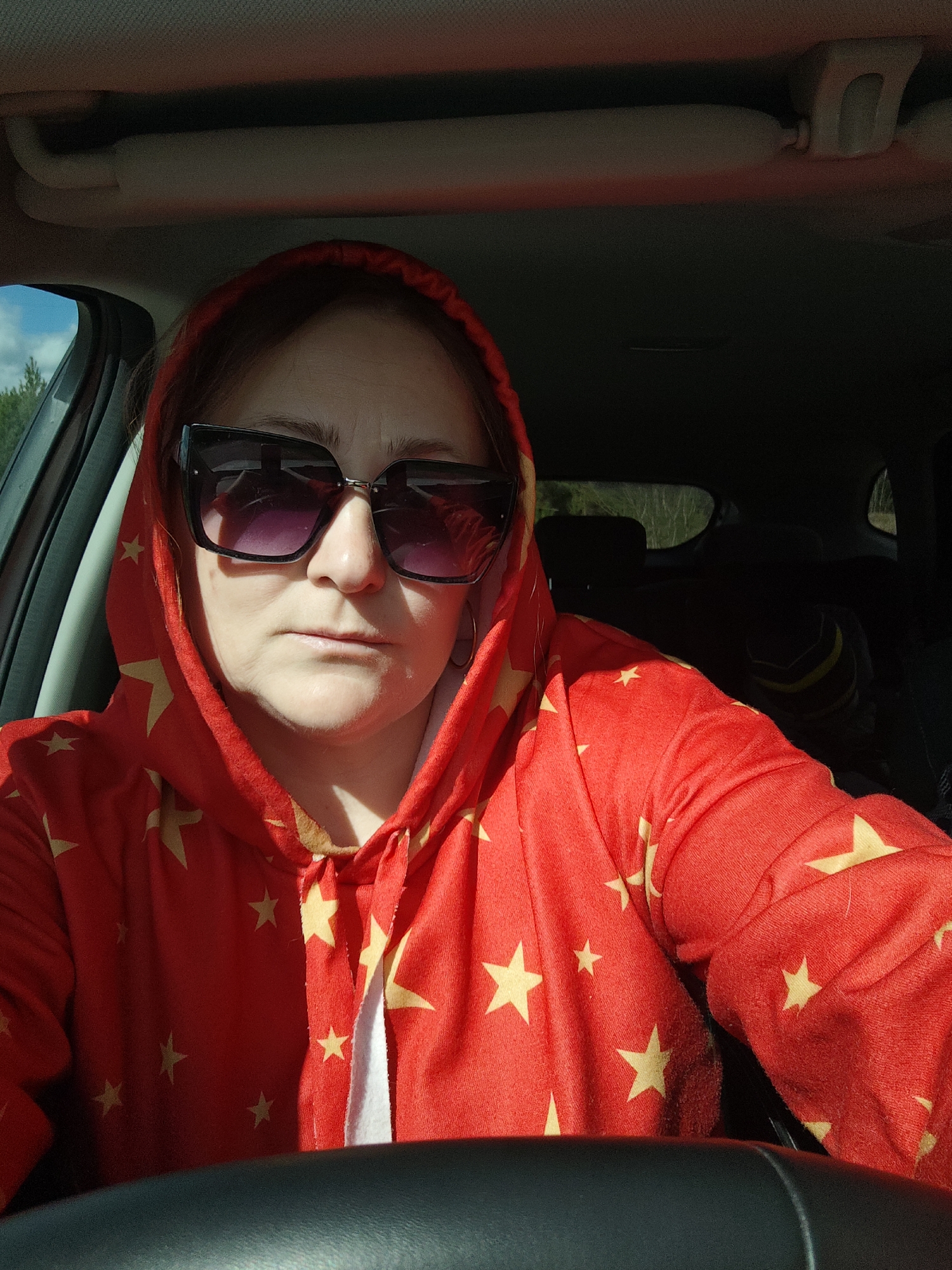 A woman is sitting in a car, wearing a red hoodie with yellow stars and large black sunglasses. She is behind the steering wheel. Trees can be seen through the window. The photograph was taken during the day. There is no evidence to suggest that this image was taken in the city of Chaikovsky, Russia.