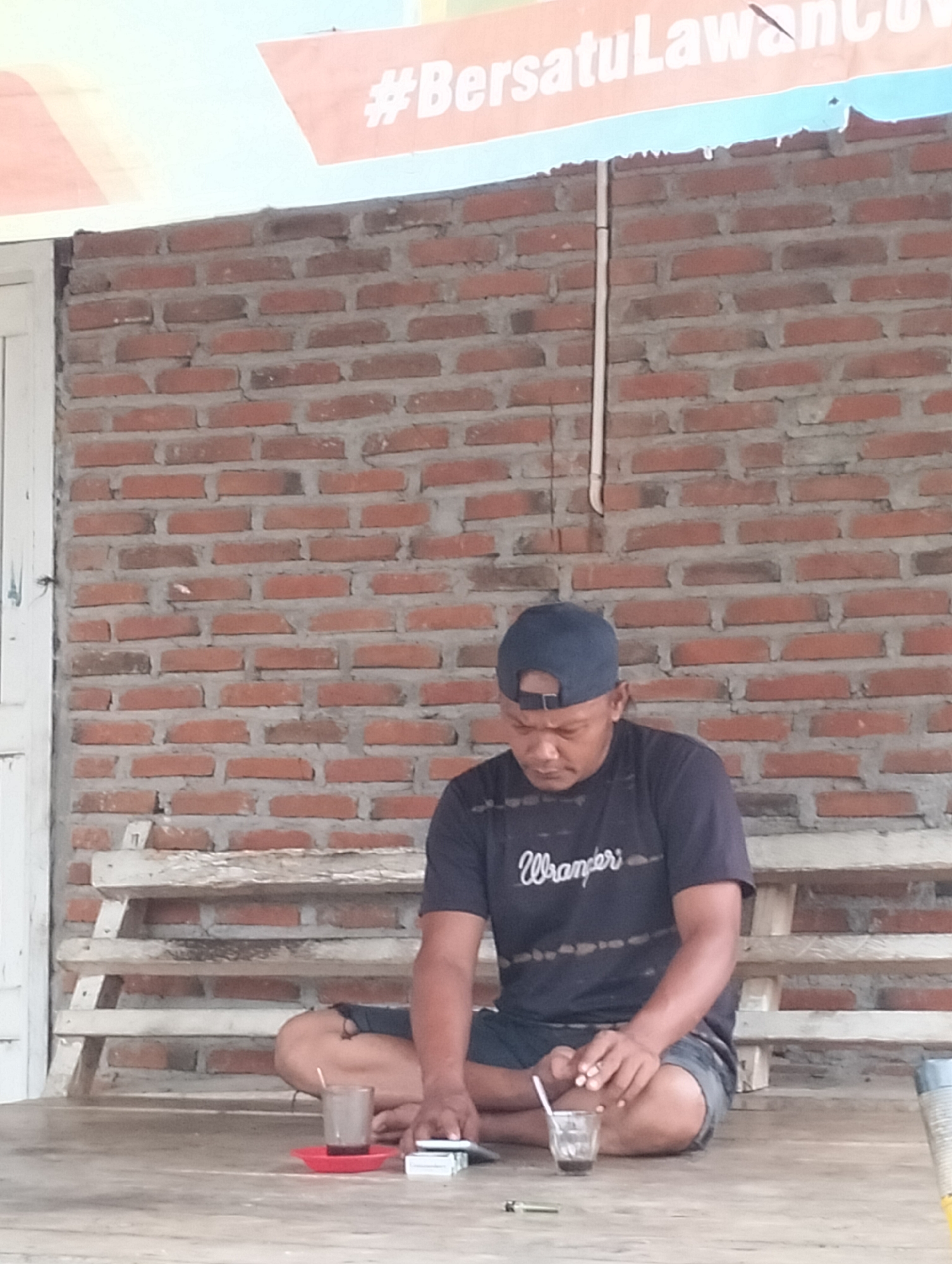A man wearing a dark blue baseball cap and a dark t-shirt with the word "Wrangler" printed on it sits cross-legged on a wooden floor in front of a brick wall. He is looking down at a small glass of dark liquid, likely coffee, with a straw, placed on a red saucer. To his right, a stack of white items, possibly cigarettes, rests on the floor. A broken wooden bench with visible slats is situated behind him against the brick wall. The brick wall shows variations in color and texture, indicating it is an outdoor or semi-outdoor structure. Above the brick wall, a banner with text in orange and blue is partially visible, suggesting a public or community area. The overall scene appears to be in a casual, rustic setting. The location is identified as Meunasahalue, Indonesia.