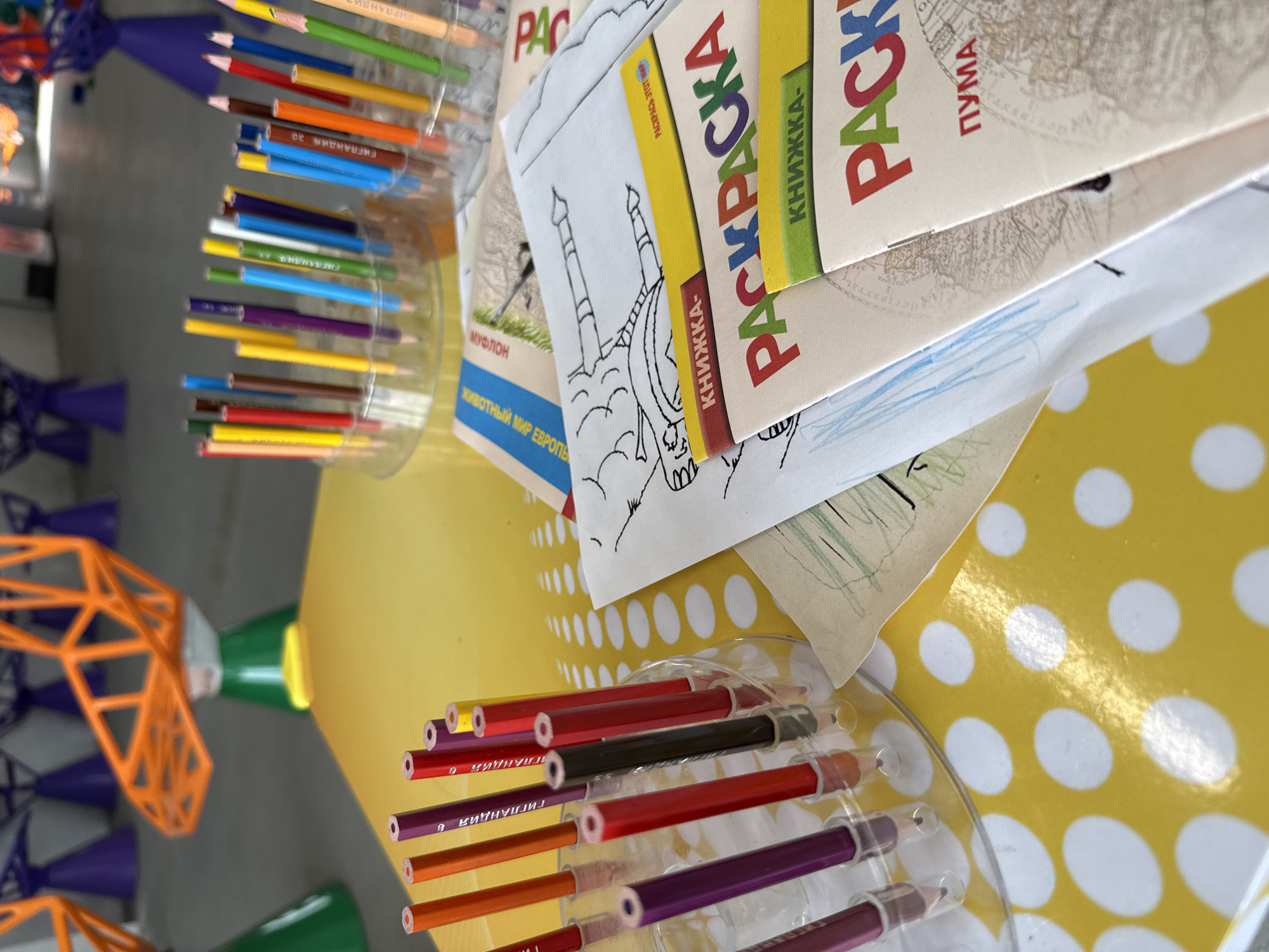 Here is a forensic description of the image:
The image presents an eye-level shot of a table in what appears to be a creative or activity-oriented space. The table is covered with a bright yellow sheet featuring white polka dots. Several coloring books titled "PACKPACHA" in Russian, stacked on top of each other, are visible. Each book displays different subtitles; "MYA," "MУФЛОН," and "ПУМА" are readable.
Two containers filled with colored pencils are placed on the table. The pencils range in colors, including shades of red, brown, yellow, purple, blue, and green.
Behind the containers, there is a construction toy.
The location in the city of Синьково, Russia is implied by the Cyrillic text on the books.