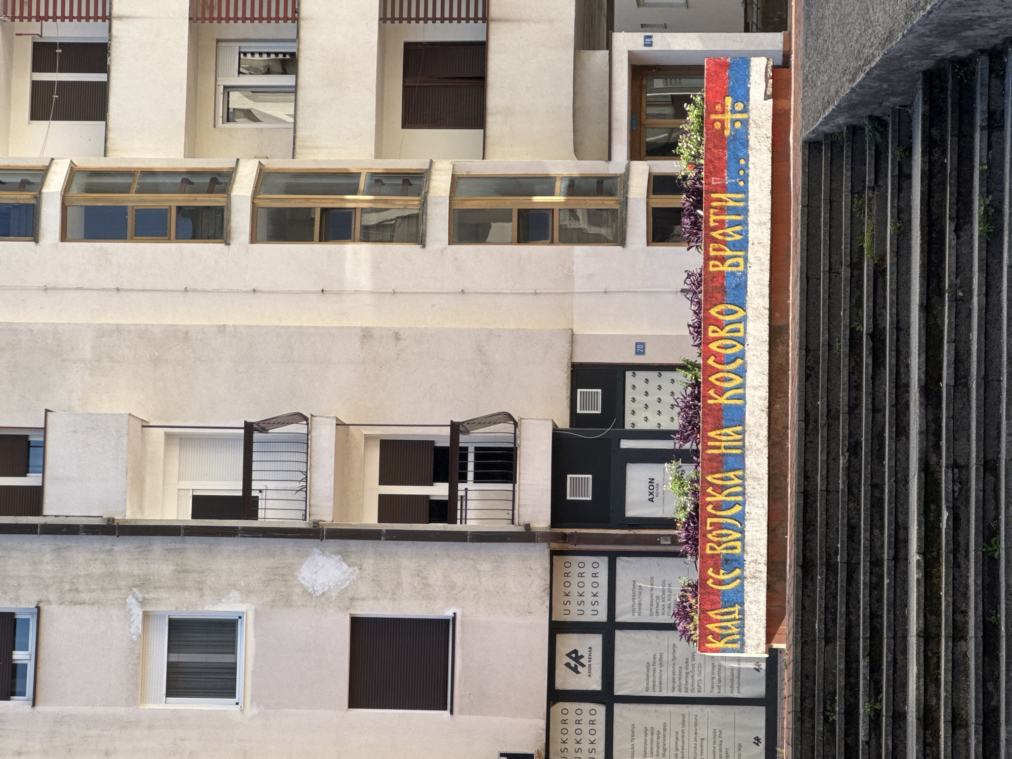 The image shows the exterior of a building in Trebinje, Bosnia and Herzegovina. The building has a modern design with multiple floors, balconies, and large windows. On the ground level, there are several shopfronts with signage, some of which are in Cyrillic script. Below these shopfronts, there is a low concrete wall adorned with a mural. The mural features the Serbian flag colors (red, blue, and white) and Cyrillic text that reads "КАД СЕ ВОЈСКА НА КОСОВО ВРАТИ..." which translates to "When the army returns to Kosovo...". A symbol, possibly related to Serbian nationalism, is also painted on the wall. In front of the building, there is a set of stone stairs leading up to the entrance. The scene is captured in daylight, with the sun casting shadows on the stairs and the building. No people are visible in the image.