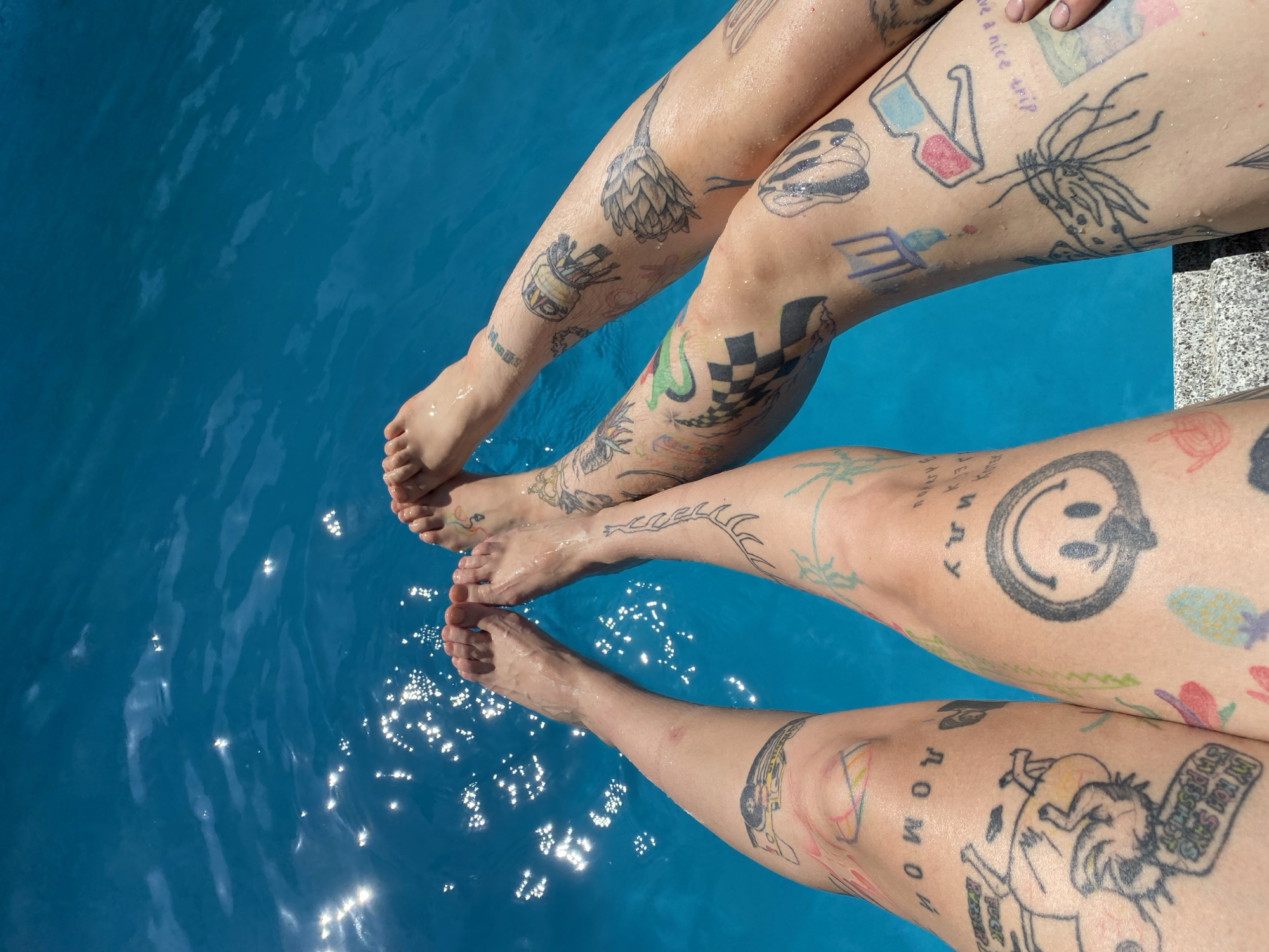This media file shows the legs of two people with numerous tattoos, dangling in bright blue water. The water is likely a swimming pool due to its clarity and the presence of a concrete edge with stone tiling on the right side of the frame. Sunlight glints off the water's surface, indicating it is daytime, likely with clear weather. The people's legs are bare and wet, suggesting they are either swimming or have just emerged from the water. The tattoos are varied, including a smiley face, a checkerboard pattern, a drawing of an artichoke, 3D glasses, a sketch of a creature, and text. Some of the visible text includes "have a nice trip" and what appears to be "AROMOM". The overall impression is one of a relaxed, summery activity, possibly at a pool or a similar water feature. The location context of Mtskheta, Georgia, does not directly influence the visual description of the image itself but might provide cultural or environmental context not discernible from the image alone.