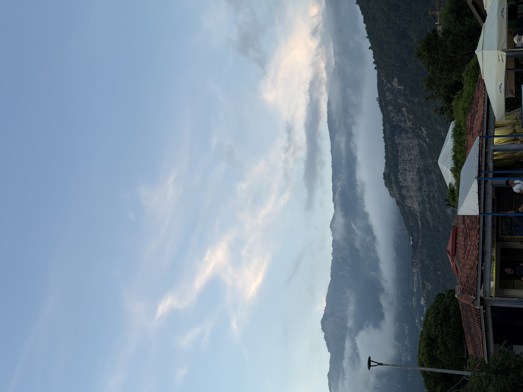 The image captures a scenic view featuring a vast mountainous landscape juxtaposed with a commercial area. The dominant feature is a long range of rugged mountains, densely covered in dark green foliage and partially obscured by atmospheric mist or low-hanging clouds that cling to their slopes. The sky above is a clear, light blue, adorned with delicate wisps of white and pale gray clouds, suggesting either late afternoon or early evening.
In the foreground, on the right side of the frame, are elements of an outdoor establishment. These include reddish-brown tiled roofs and several white awnings or large umbrellas, some bearing visible text. The words "ICE CREAM" are clearly displayed on multiple awnings, and the brand name "Frikoma" is also visible on one, indicating an ice cream parlor or a similar vendor. A few indistinct individuals are visible near these structures, including a person in a white t-shirt and another in a dark t-shirt. A black street lamp post stands towards the lower left. The overall impression is one of tranquility and natural beauty, characteristic of a coastal or mountainous region like Budva, Montenegro, where such a blend of scenic vistas and local amenities is common.