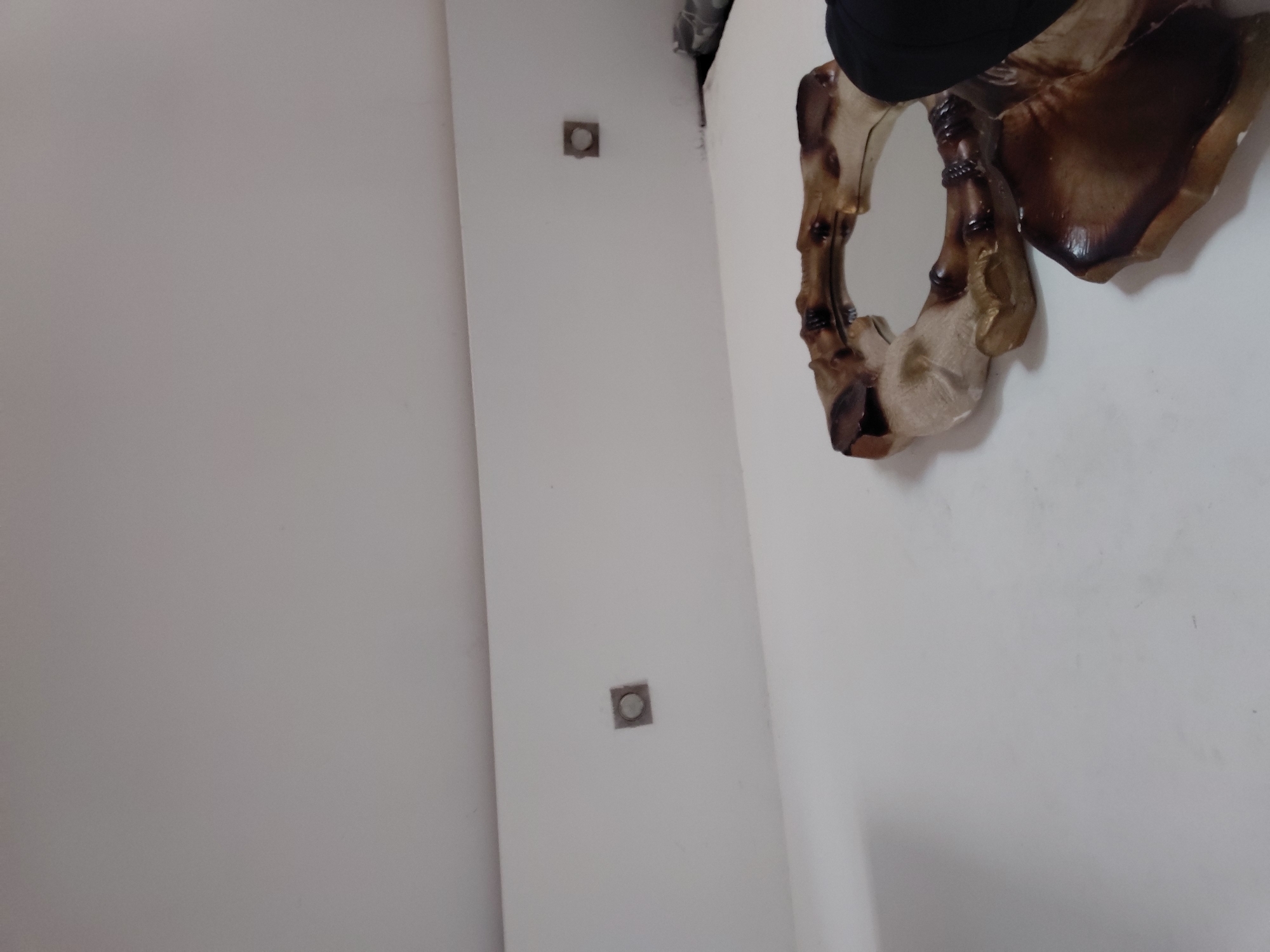 The image displays an indoor scene, primarily featuring a white wall. On the right side, a decorative wall mirror with a distinctive, organic-shaped frame is partially visible. The frame appears to be crafted from wood or a similar material, exhibiting various shades of brown and beige, with darker, burnished-like accents and a somewhat rustic or natural texture. A portion of a black object is visible at the very top edge, seemingly located above the mirror. On the left side of the white wall, two small, square, metallic-grey fixtures are mounted, each containing a central circular indentation or button. These fixtures are placed vertically along the wall. The photograph is taken from a slightly low and angled perspective, making the wall appear to recede diagonally. There is no specific activity taking place, and no people are present. The lighting is even, suggesting an indoor setting without strong natural light sources. No text is visible within the image.