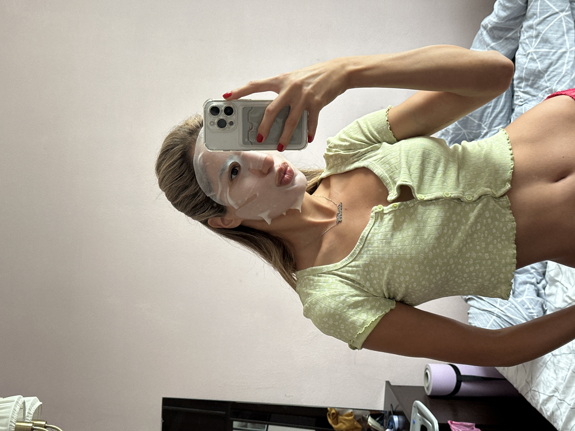 The image shows a young woman with a sheet mask on her face, taking a selfie with her phone. She is wearing a light green, short-sleeved cropped top with a floral pattern. Her hair is long and blonde. She is leaning back, with her right arm extended to hold the phone. The background includes a bed with blue and white bedding, and a pink yoga mat on a dark surface. The overall lighting suggests it is daytime indoors. The woman's expression is playful, with her lips pursed. There is no visible text in the image. The context provided indicates the location is Moscow, Russia.