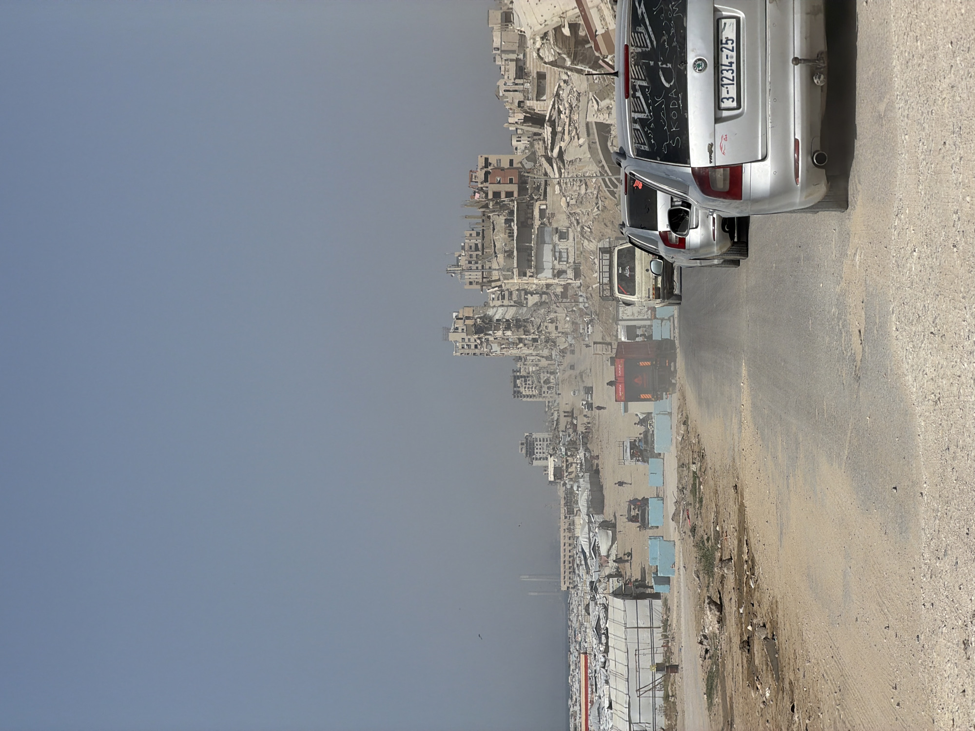 The image shows a scene of destruction, possibly after a conflict or natural disaster. Buildings in the distance are heavily damaged, reduced to rubble and skeletal structures. Several vehicles, including cars and what appears to be a fire truck, are on a road. The road itself is dusty and possibly damaged. The sky is a pale blue, suggesting either a hazy or polluted atmosphere. The foreground shows a roadside with scattered debris and temporary structures, possibly refugee camps. The cars in the foreground are closest to the viewer, one has Arabic writing on the back windshield. The license plate on the vehicle is a white rectangle with the numbers '3-1234-25' printed in black. The location is a city in Unknown, Unknown.