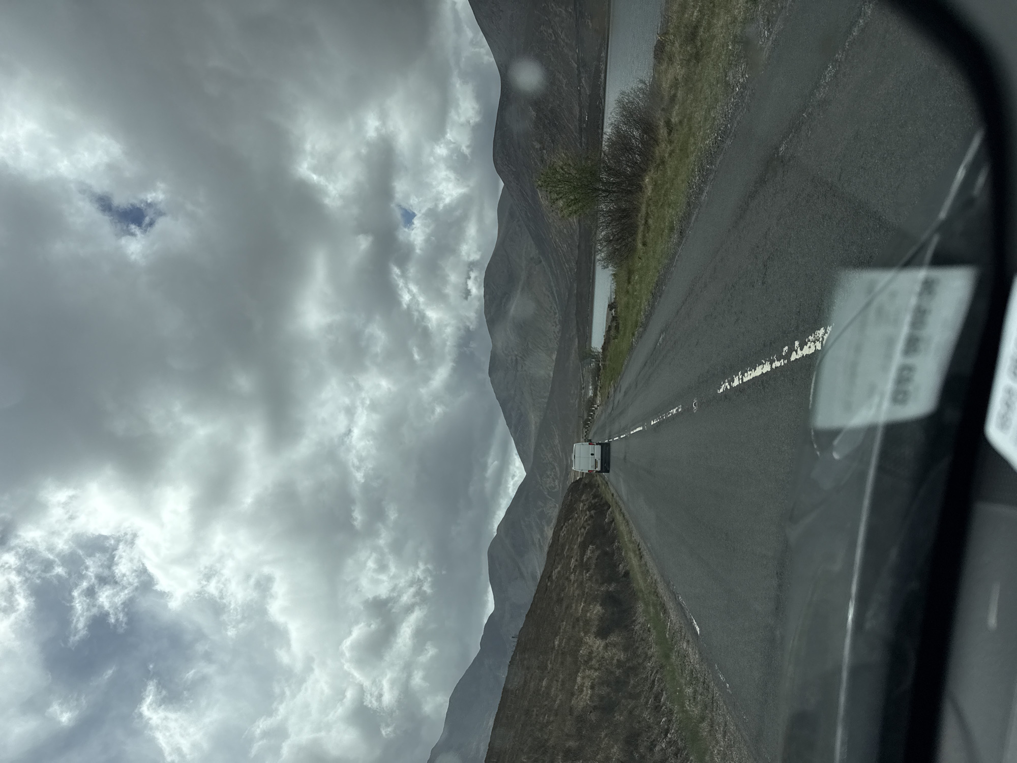 The image depicts a gray asphalt road winding through a mountainous landscape under a cloudy sky. A white van is visible on the road. Adjacent to the road is a narrow strip of vegetation along the edge of a body of water. The image is taken from inside a vehicle, with a partial view of the vehicle's interior visible in the lower right corner. The location is unknown.