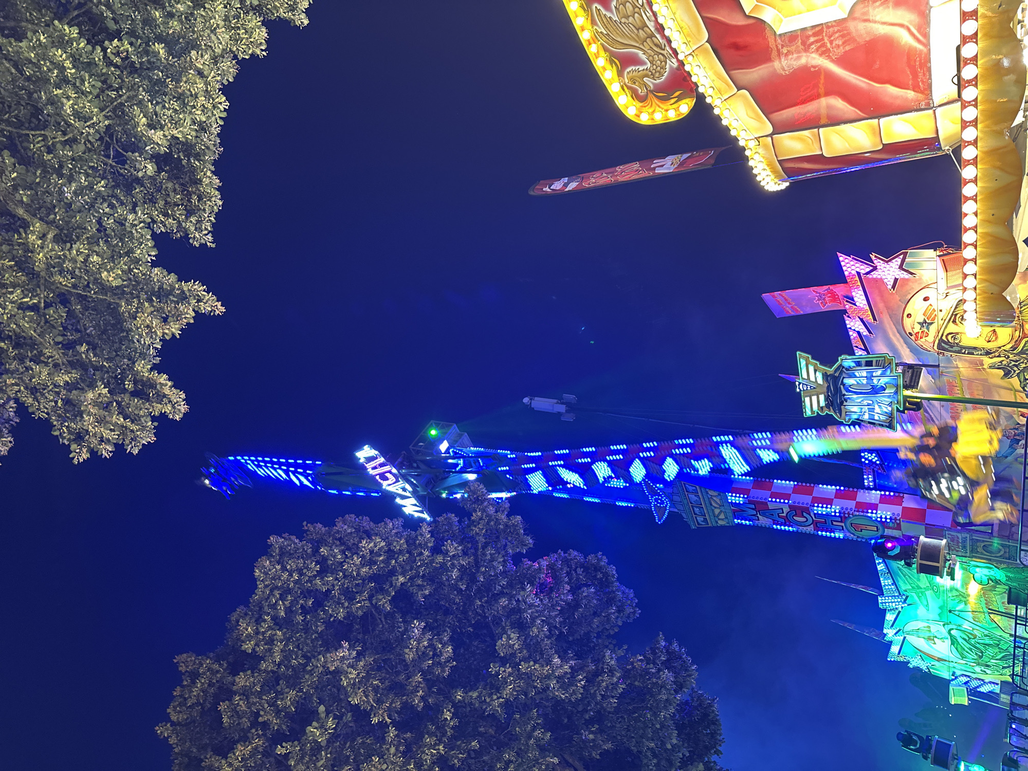 The image captures a nighttime scene at a funfair in Bruchhausen-Vilsen, Germany. Dominating the frame is a tall, brightly lit amusement ride with vertical blue and white lights ascending its structure. The word "MACHI" is illuminated in blue letters at the top of the ride. Below this, a checkered pattern of red and white squares is visible. Several gondolas are attached to the ride, with some appearing to be in motion, indicated by blur. To the left of the main ride, trees with foliage are silhouetted against the dark blue night sky. In the foreground to the right, part of another attraction is visible, featuring a red surface adorned with a golden eagle motif and outlined with numerous small, yellow lights. Further to the left, another amusement ride element is present, illuminated with green and pink lights, and depicting a stylized image of a person wearing a helmet. The overall atmosphere is one of vibrant nighttime entertainment.