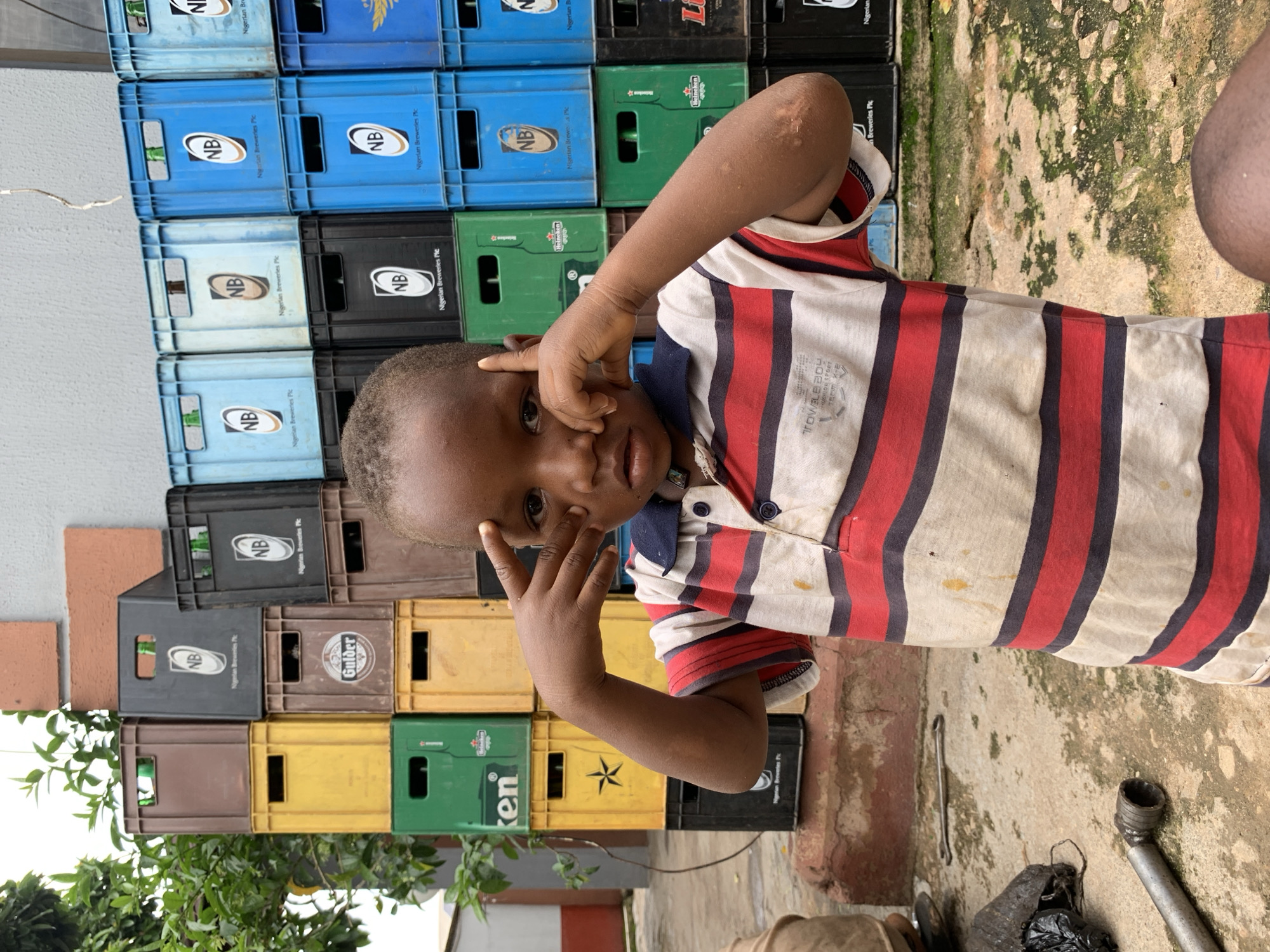 The image depicts a young boy standing in front of stacked crates of beverages in Jos, Nigeria. The boy, wearing a striped polo shirt, looks directly at the camera with his fingers touching his temples. Behind him, a wall of crates, predominantly blue and brown, are neatly stacked. Some of the crates bear branding, including "NB" and "Nigerian Breweries Plc". The ground is a mix of concrete and dirt, with scattered debris including a metal tool, possibly a wrench, and a black plastic bag. Lush green foliage is visible in the upper left corner of the frame. The overall scene suggests an outdoor storage area, possibly behind a business or a residential compound.