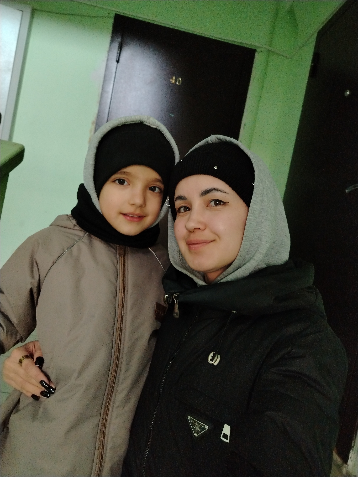 This indoor portrait features two individuals, likely a mother and child, posing for a photograph in what appears to be a hallway or corridor in Noviy, Russia. Both are dressed in warm attire, including hooded jackets and beanies, suggesting a cool climate.
The adult, positioned on the right, wears a black beanie with small decorative studs, a grey hooded sweatshirt, and a black puffer jacket. She has a subtle, content smile, a nose piercing, and dark nail polish on her left hand, which also bears rings. The child, on the left, wears a black beanie, a black neck gaiter, and a light brown or tan hooded jacket. The child looks directly at the camera with bright, engaging eyes and a soft, slight smile.
The background consists of light green painted walls and two dark doors. The number "40" is visibly affixed to the door behind the child. A portion of a white doorframe or window is visible on the far left. Notable details on the adult's jacket include a small, silver-colored emblem and a triangular patch with the text "FASHION." The overall scene conveys a casual and familial moment.