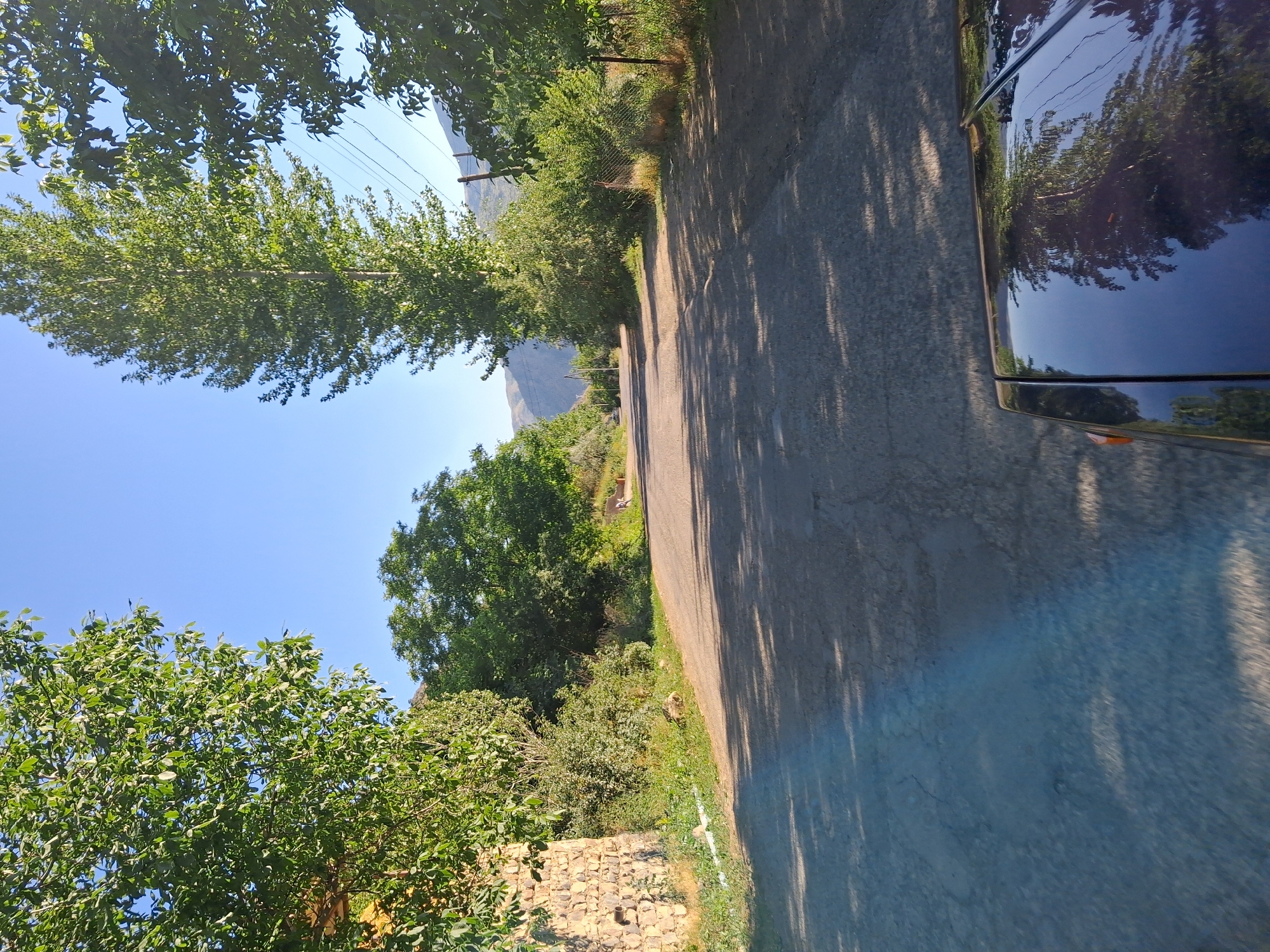 This media file shows a view from a vehicle driving on a paved road through a rural area. The scene is dominated by lush green trees and vegetation, with a clear blue sky above. The road is shaded by trees, casting dappled shadows on the asphalt. On the right side of the image, the side and roof of a dark-colored vehicle are visible, reflecting the surrounding trees and sky.
In the background, mountains can be seen in the distance under the bright sky. The overall impression is of a sunny day in a picturesque, natural setting. The time of day appears to be morning or afternoon, given the angle of the sun and the presence of strong shadows. There are no people or explicit text visible in the image. The setting suggests a drive through a countryside location, possibly in a mountainous region, consistent with the provided location context of Shatin, Armenia. The image captures a serene and tranquil moment of travel through a green landscape.